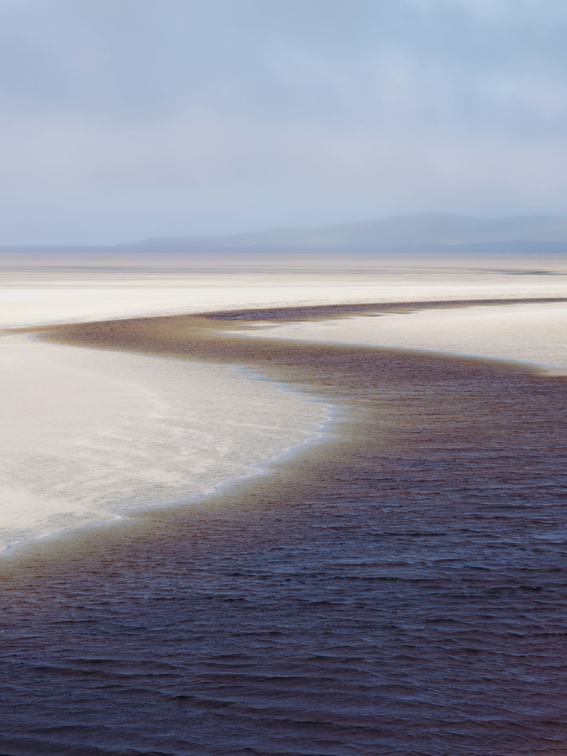 Dark water winds through a pale, sandy expanse under a cloudy sky on the Hebrides Islands, Scotland.