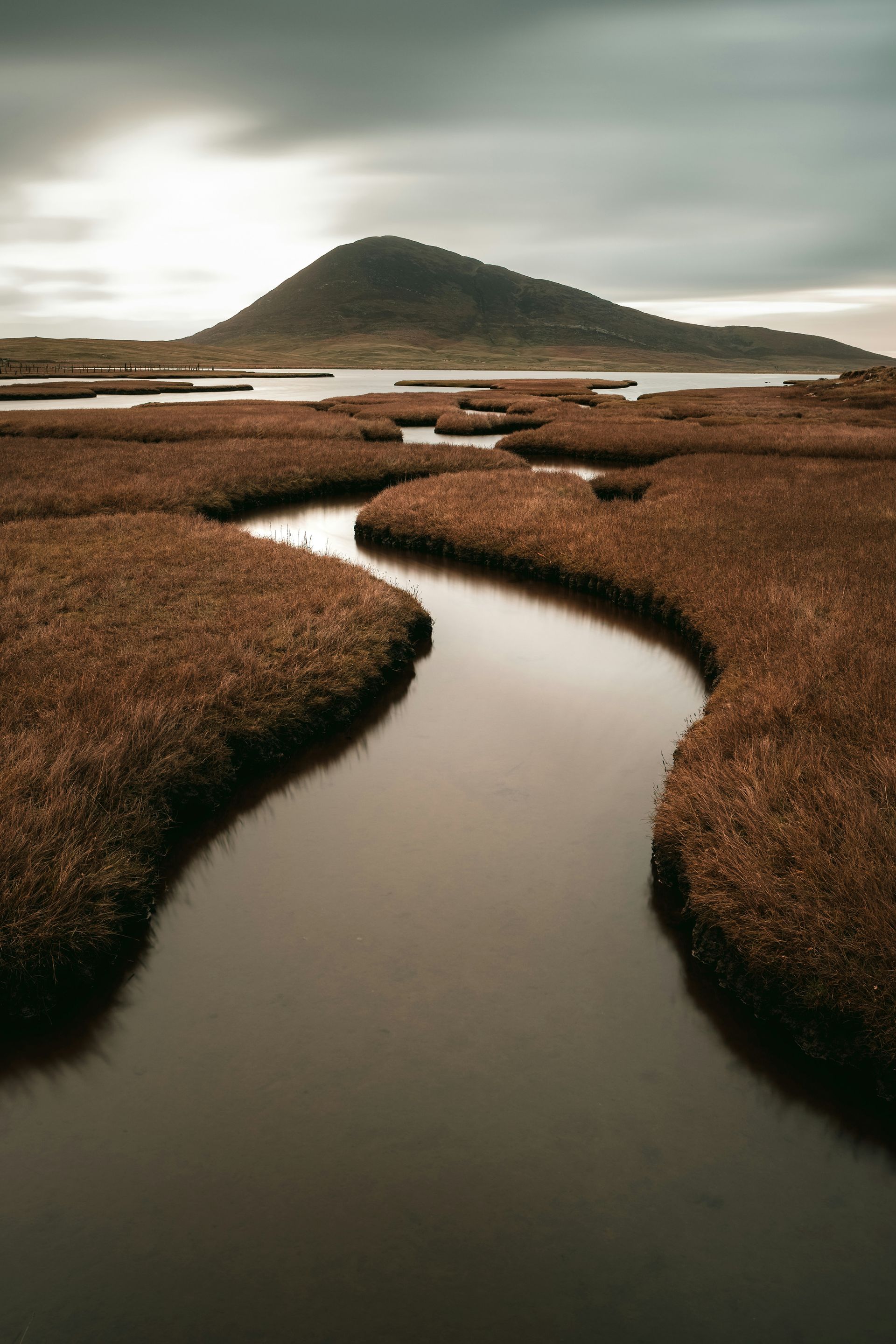 A winding river through brown marshland, with a dark hill in the background on the Hebrides Islands, Scotland.