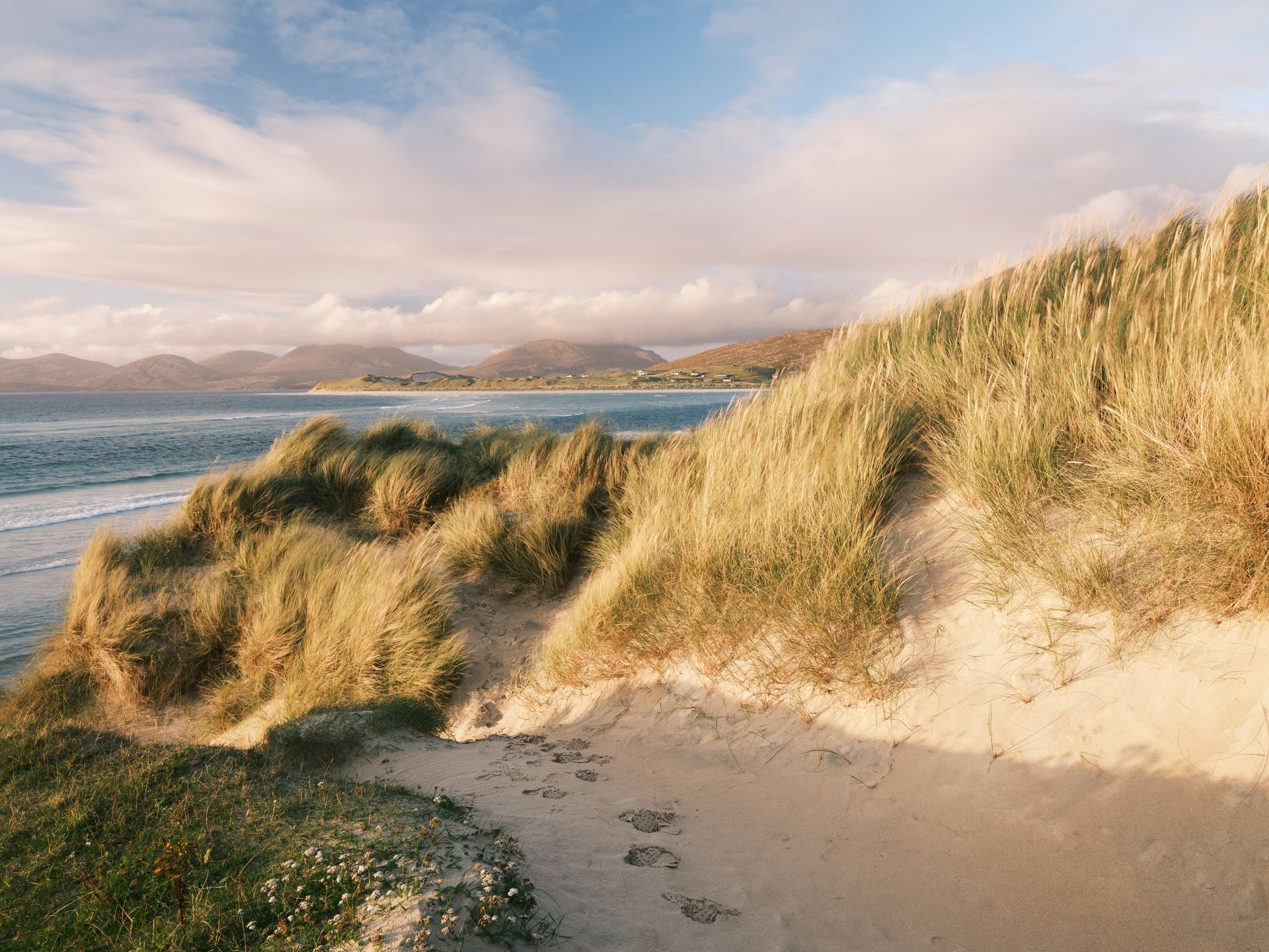 Sandy dunes with tall grass overlook a beach and ocean under a partly cloudy sky on the Hebrides Islands, Scotland.