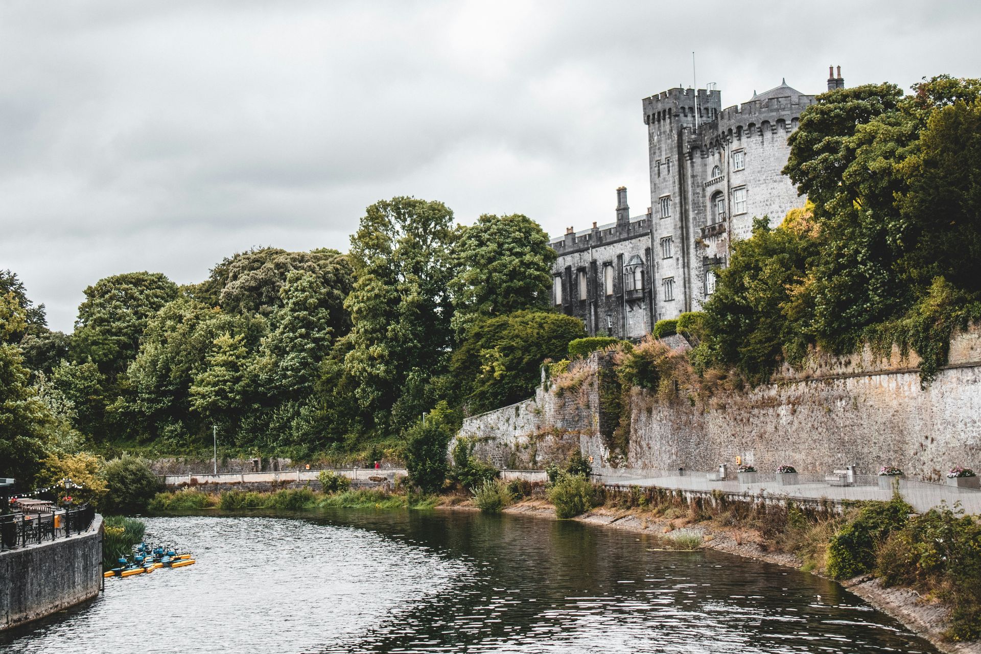 Kilkenny Castle, Ireland, overlooks the River Nore, partially visible. Stone walls, trees, and a cloudy sky.
