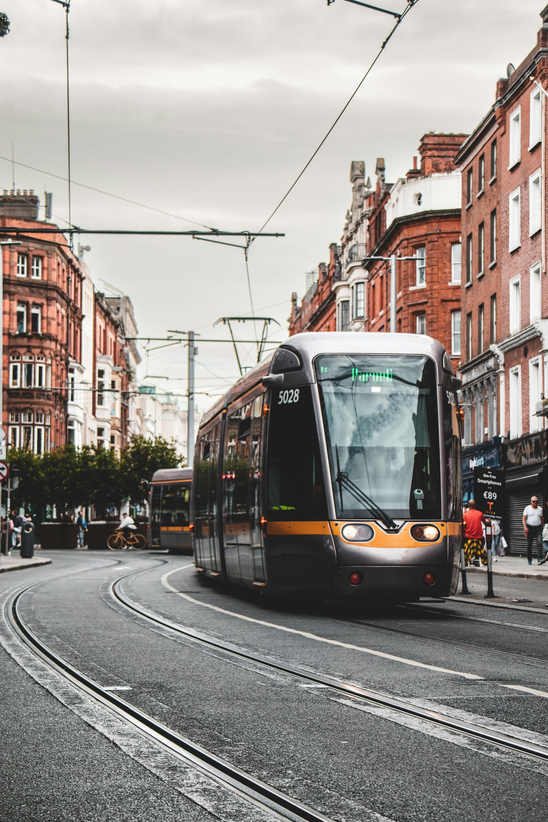 Tram on tracks in a city street, passing buildings with red brick facades, under a cloudy sky in Kilkenny, Ireland.