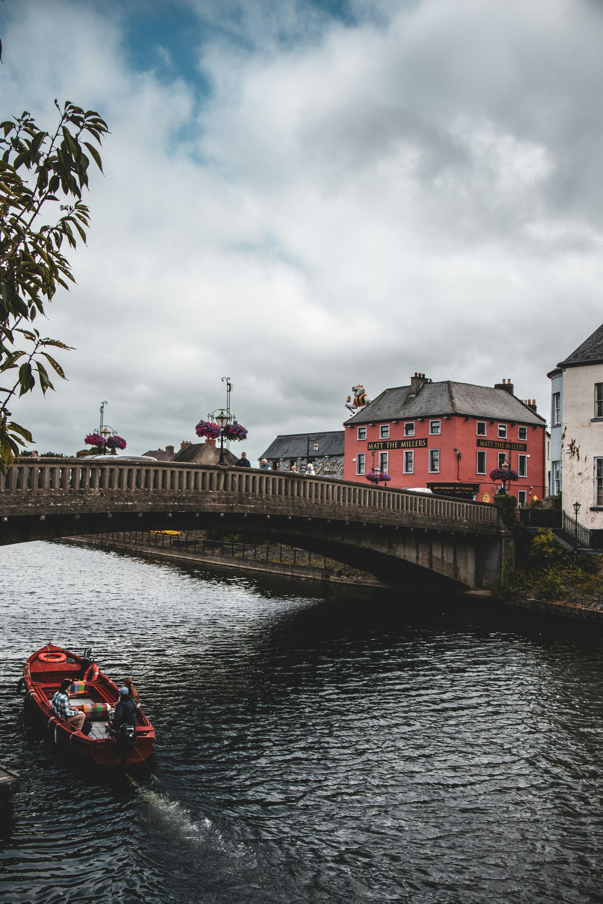 A red boat on dark water, a stone bridge, and a pink building under a cloudy sky in Kilkenny, Ireland.