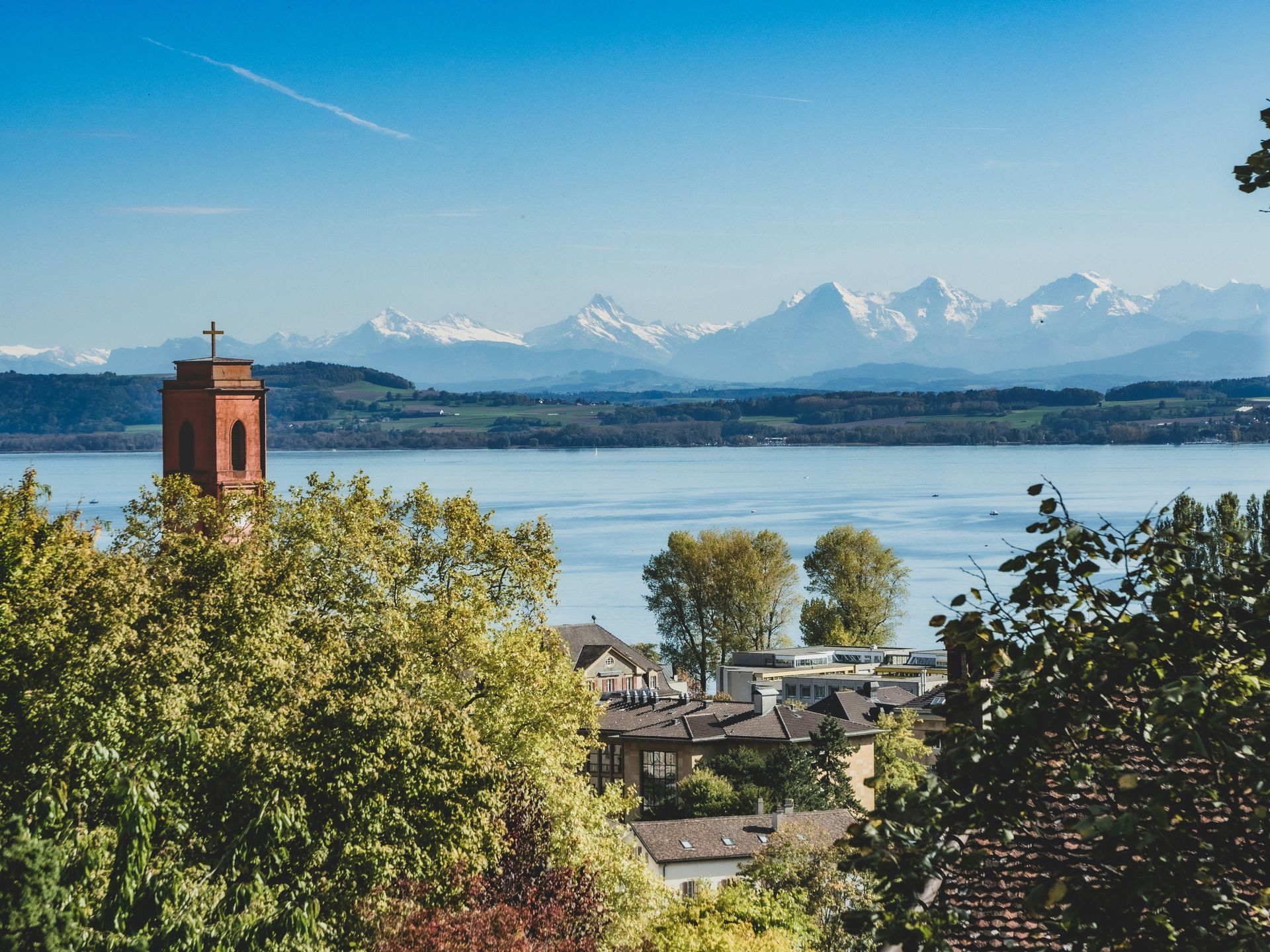 A lake scene with a church tower, houses, trees, and snow-capped mountains under a blue sky in Neuchâtel, Switzerland.
