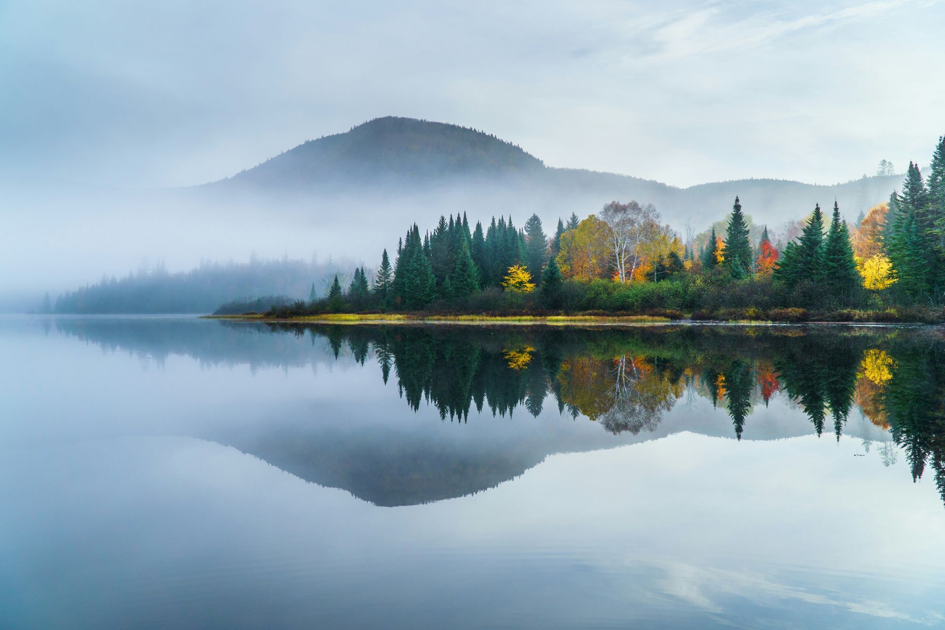 A misty lake reflects a forested shoreline and a mountain peak. Autumn colors dot the trees in Canada.
