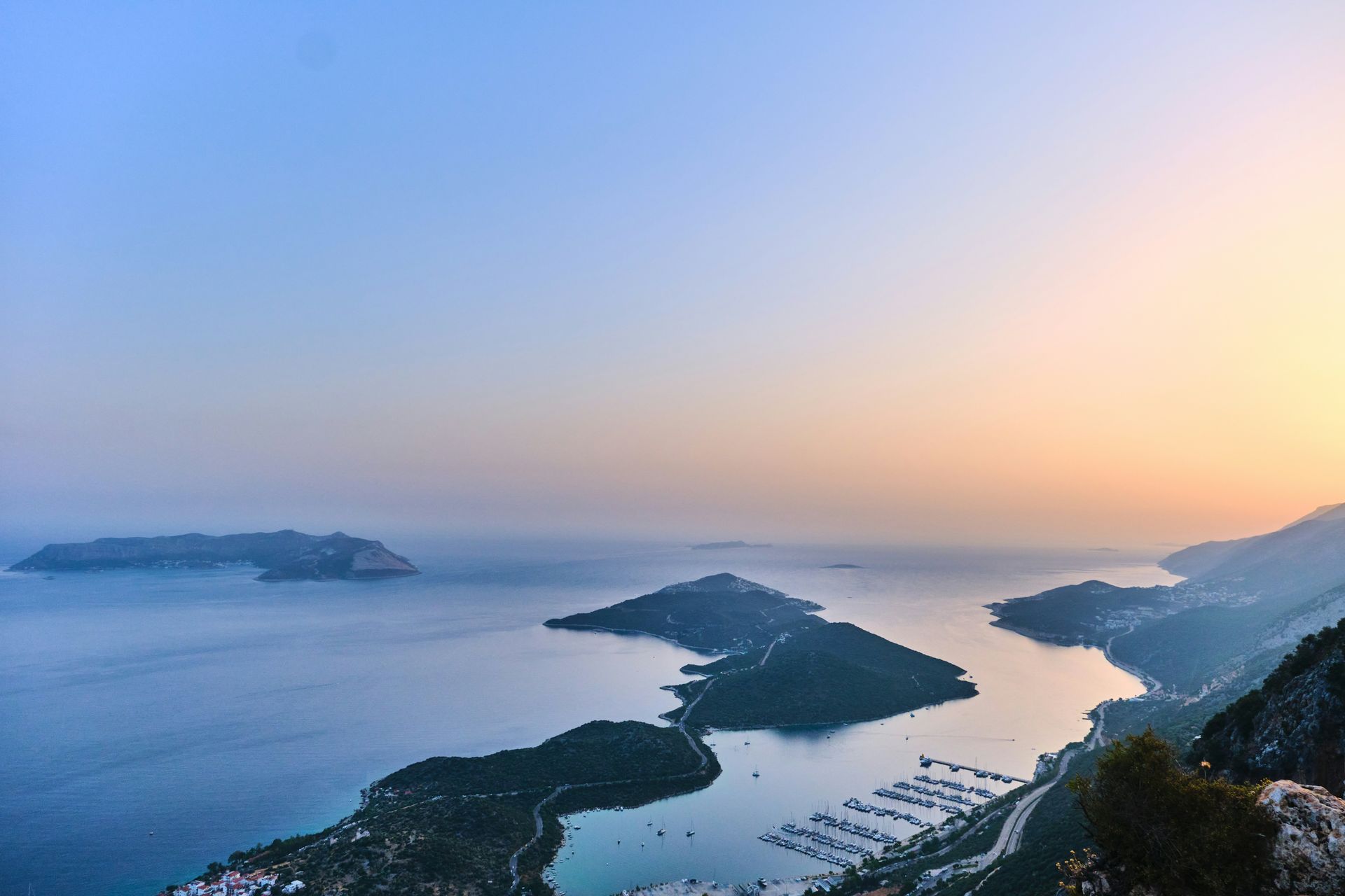 Coastal landscape at sunset: islands dot the calm blue sea, with a marina nestled along the shoreline in Kaş, Turkey.