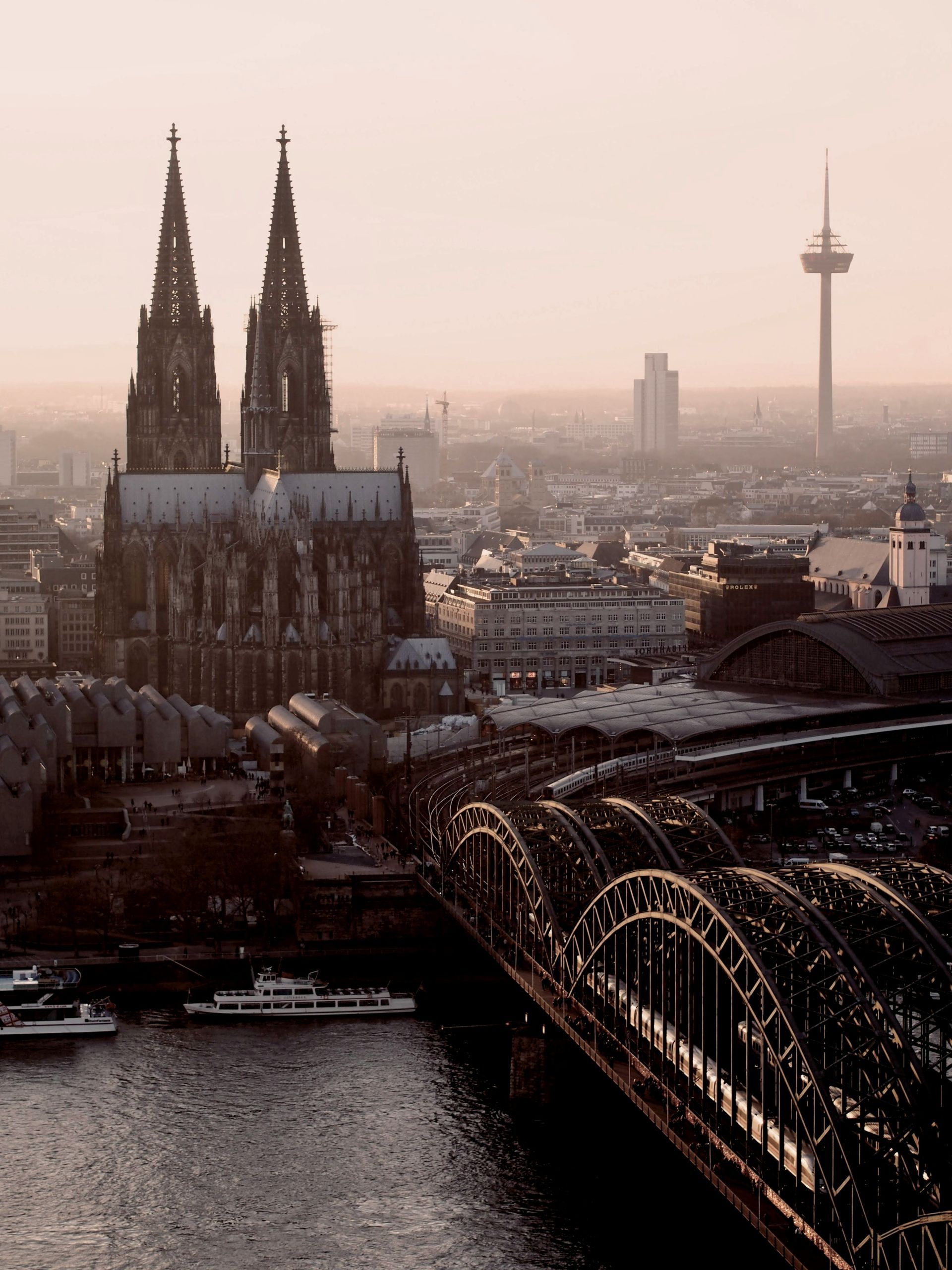 Cologne skyline at sunset: Cathedral, bridge over river, and telecommunications tower in soft light.