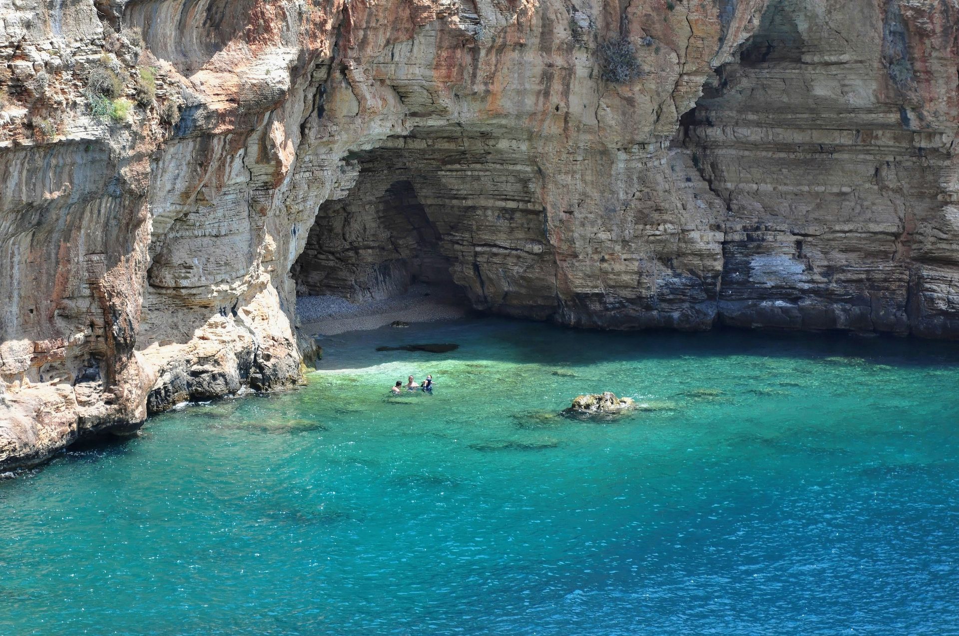 Cave opening on a rocky cliffside, leading to turquoise water. Several people and boats in Antalya, Turkey.