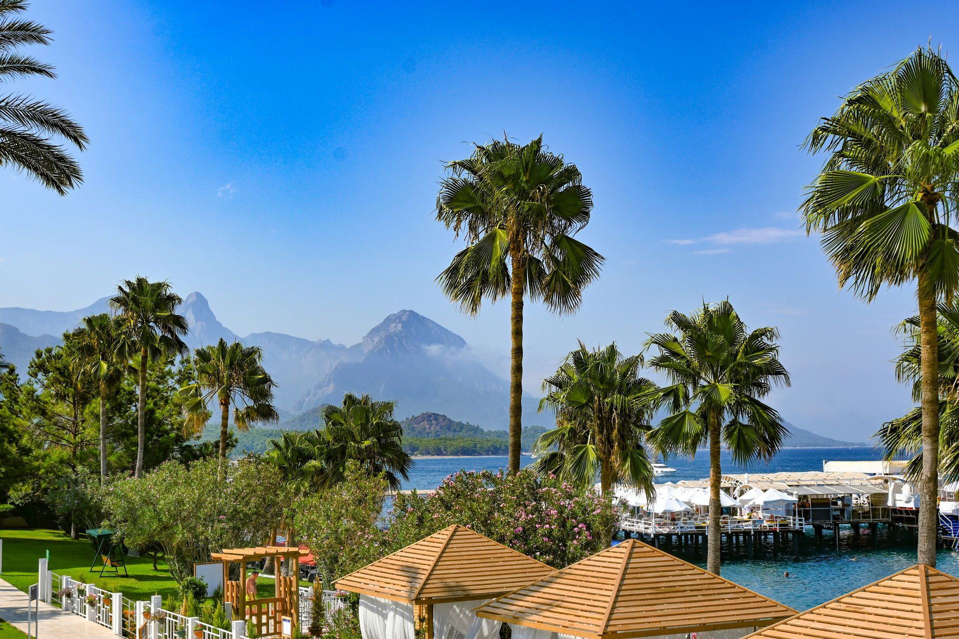 Palm trees frame a coastal view: mountains, blue sea, and a white pier in Antalya, Turkey.