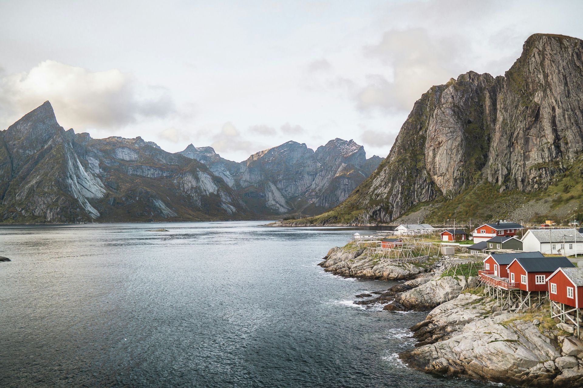 Red cabins on the shore of a lake with jagged mountains under a cloudy sky in Lofoten, Norway.