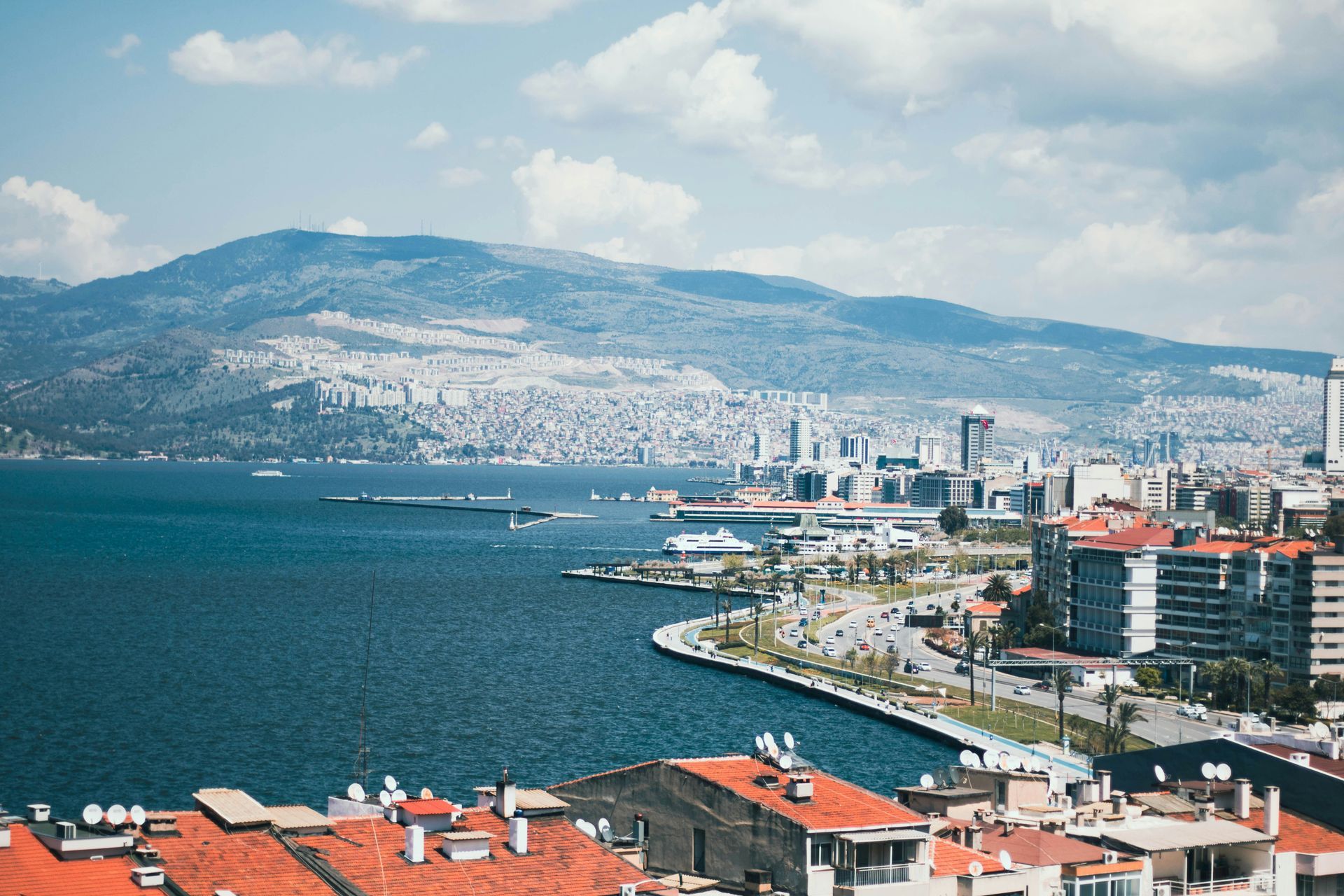 Coastal city view of İzmir, Turkey, with buildings, sea, and a mountain under a partly cloudy sky.