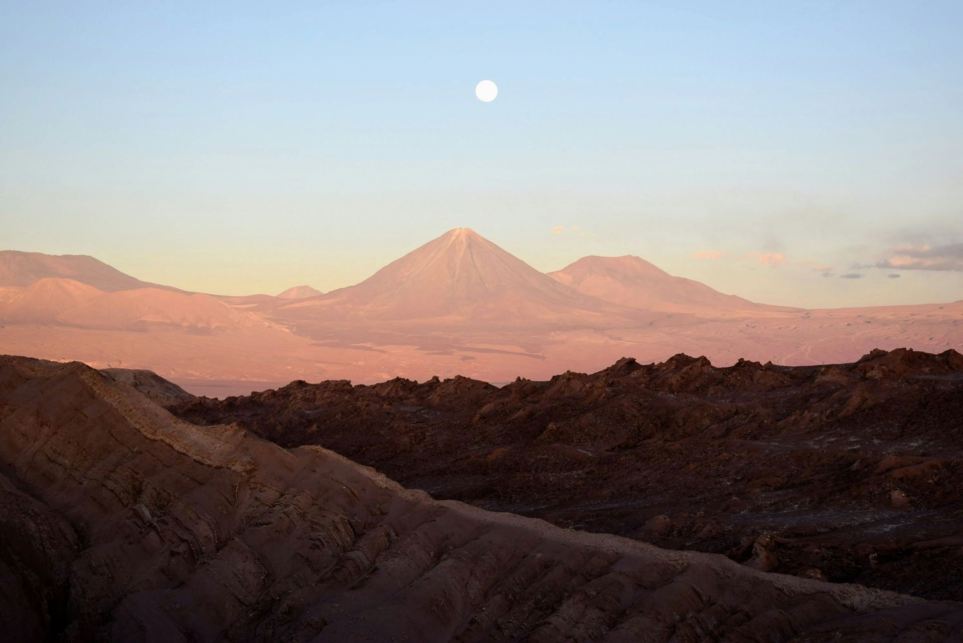 Moonlit mountain range at dawn, pink and tan hues over barren terrain in the Atacama Desert, Chile.