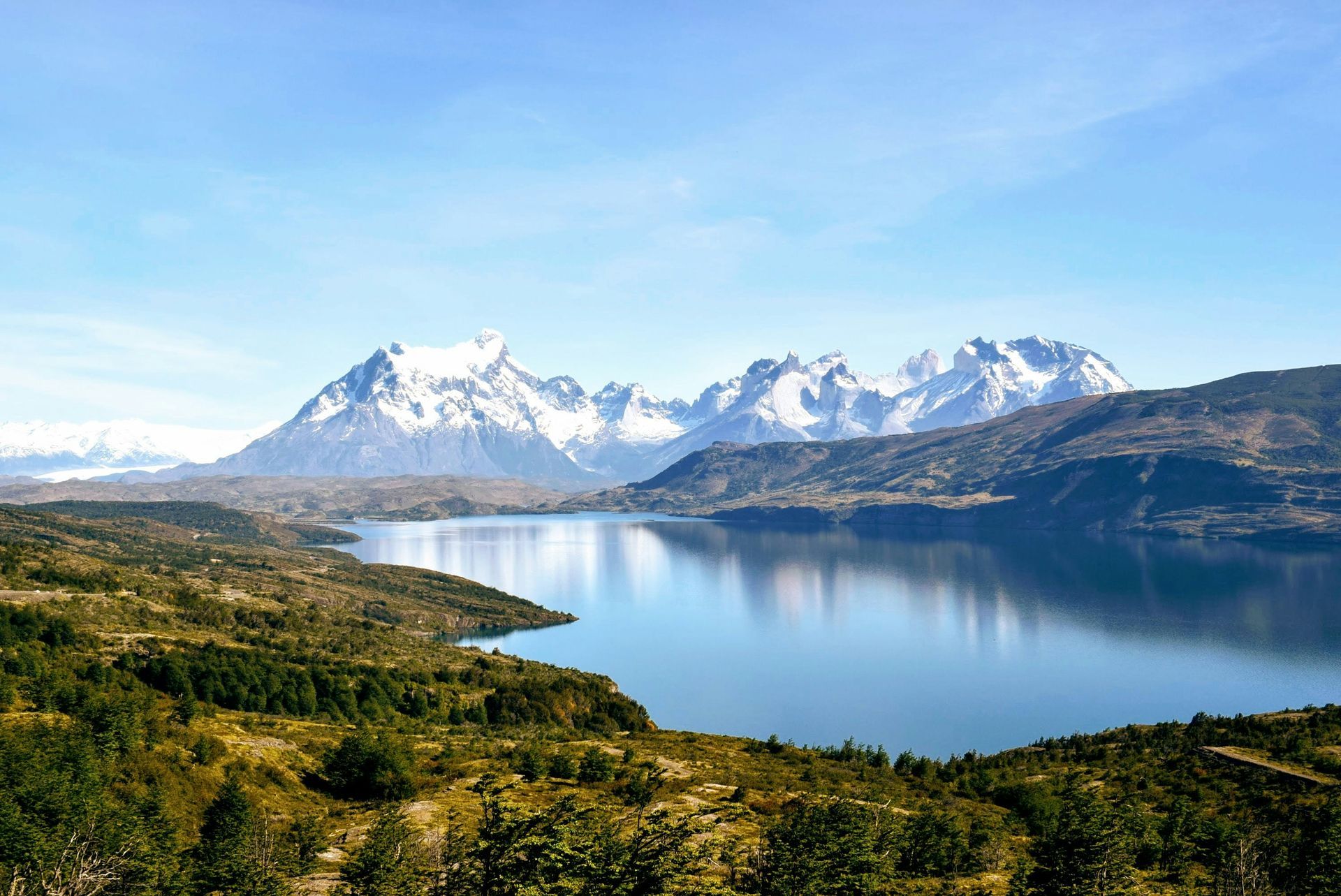Snow-capped mountains reflect in a calm blue lake, with green and brown vegetation in the foreground in Torres del Paine, Chile.