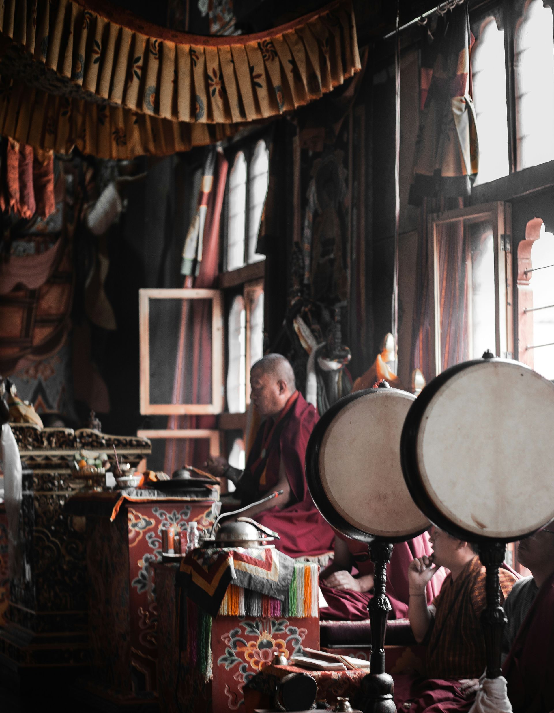 Buddhist monk playing drums in a temple, surrounded by colorful items and windows in Thimphu, Bhutan.