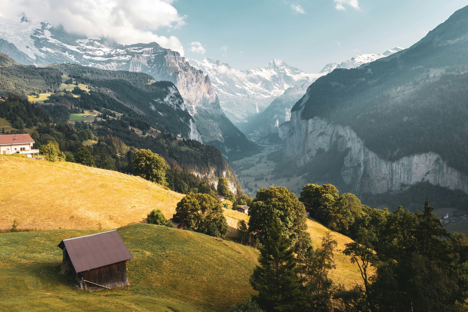 Rolling green hills and mountains in a valley, with a cabin and buildings in the foreground. Snow on peaks in Lauterbrunnen.