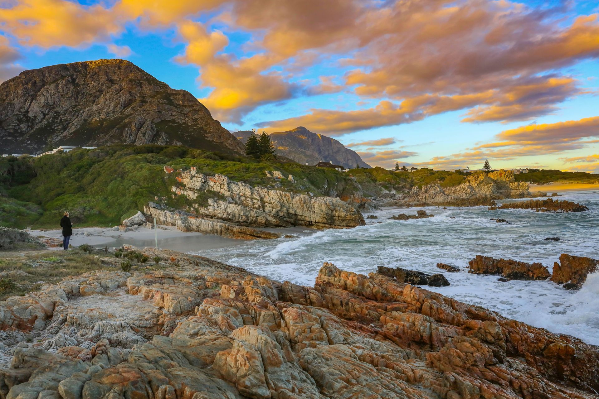 Rocky coastline with mountains under a sunset sky; a person stands on the beach in Hermanus, South Africa.