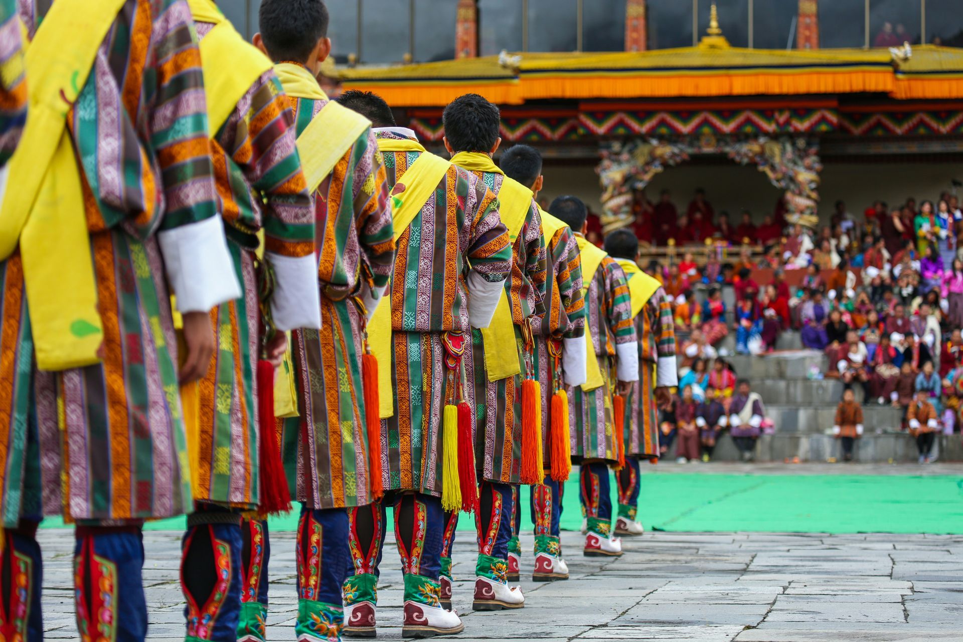 Men in colorful traditional Bhutanese clothing lined up, facing away, performing outdoors in Thimphu, Bhutan.