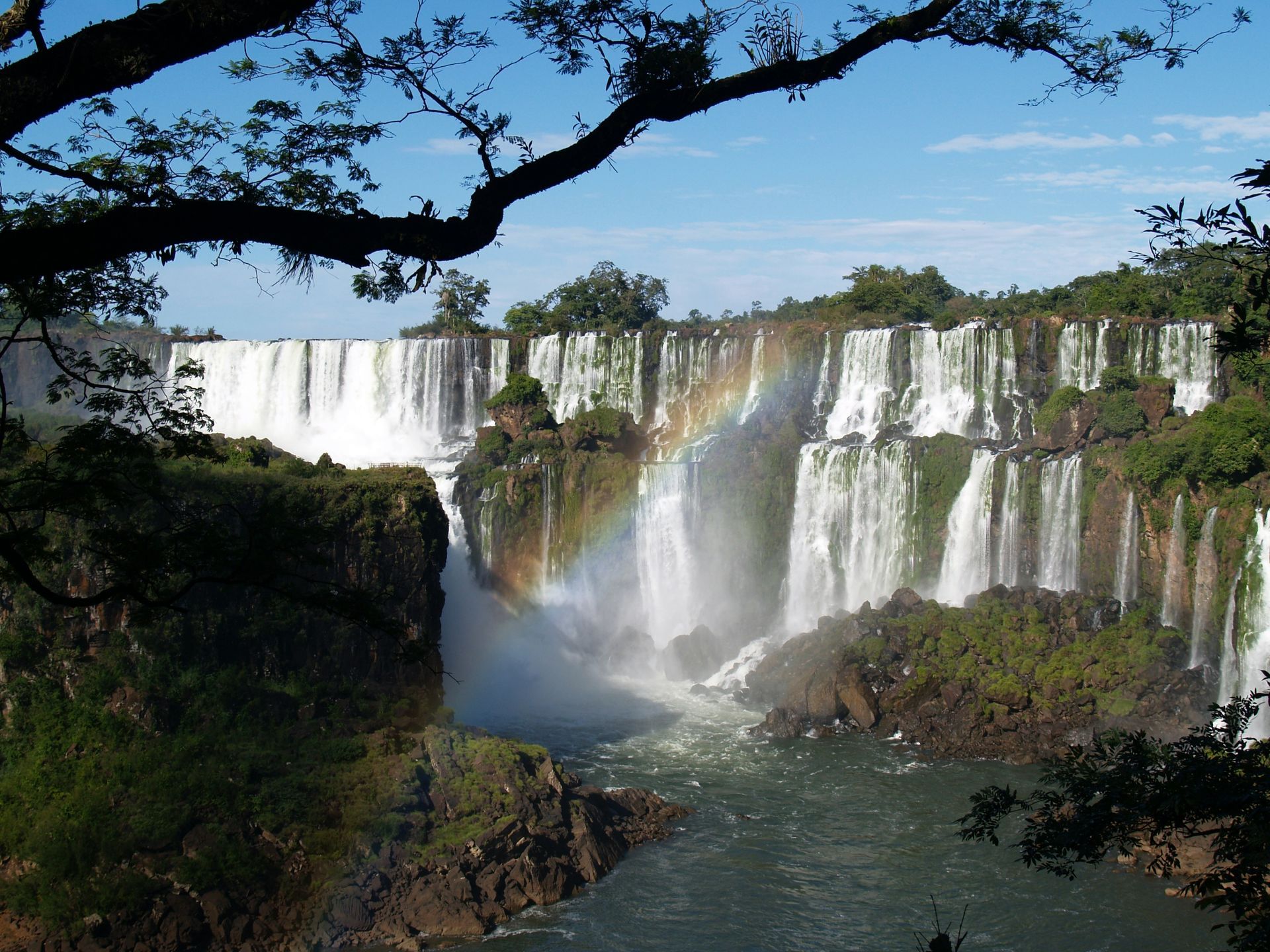 Waterfall with rainbow, lush green foliage, and a bright blue sky at Iguazu Falls, Argentina.