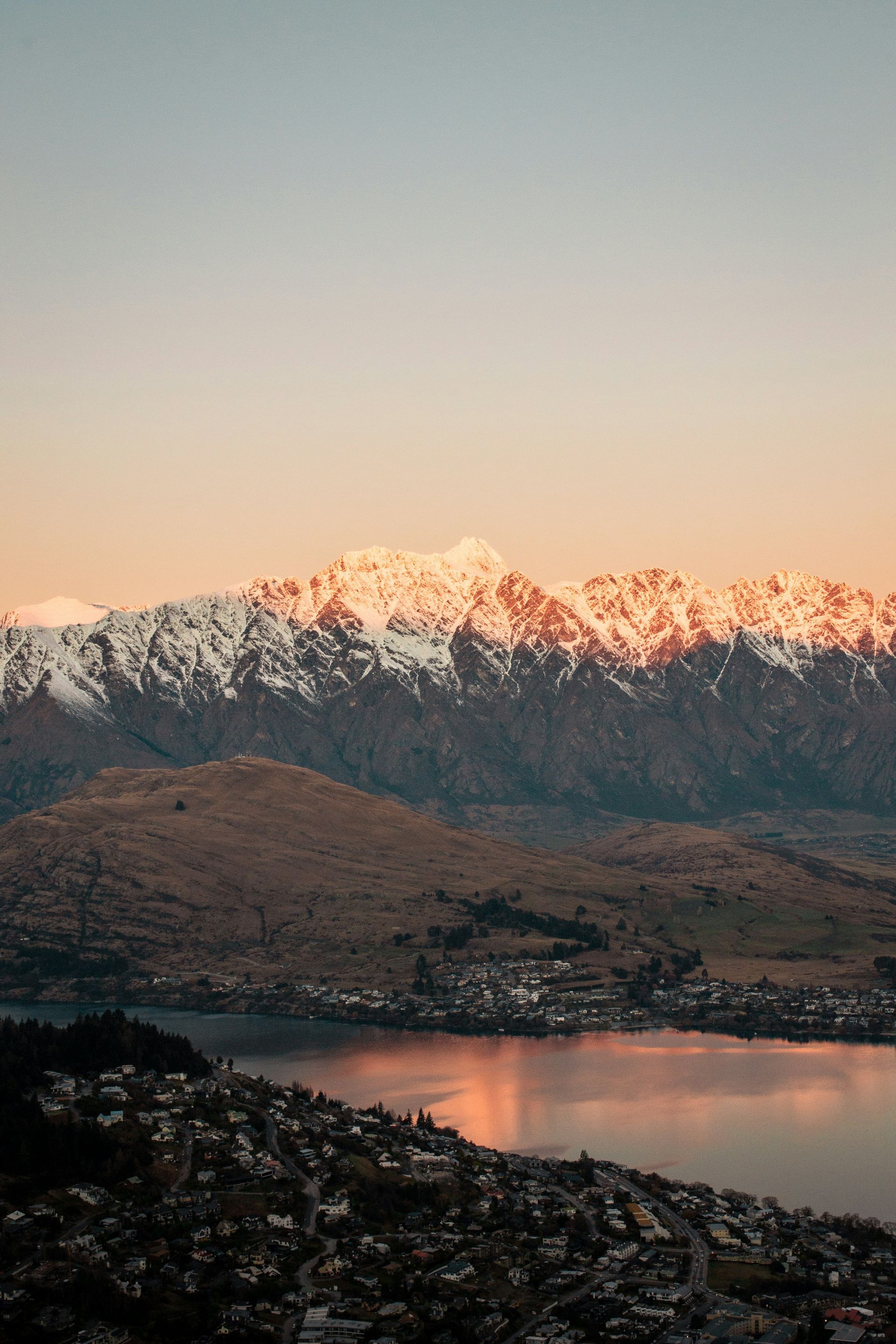 Queenstown, New Zealand is surrounded by snow covered mountains and a lake at sunset.