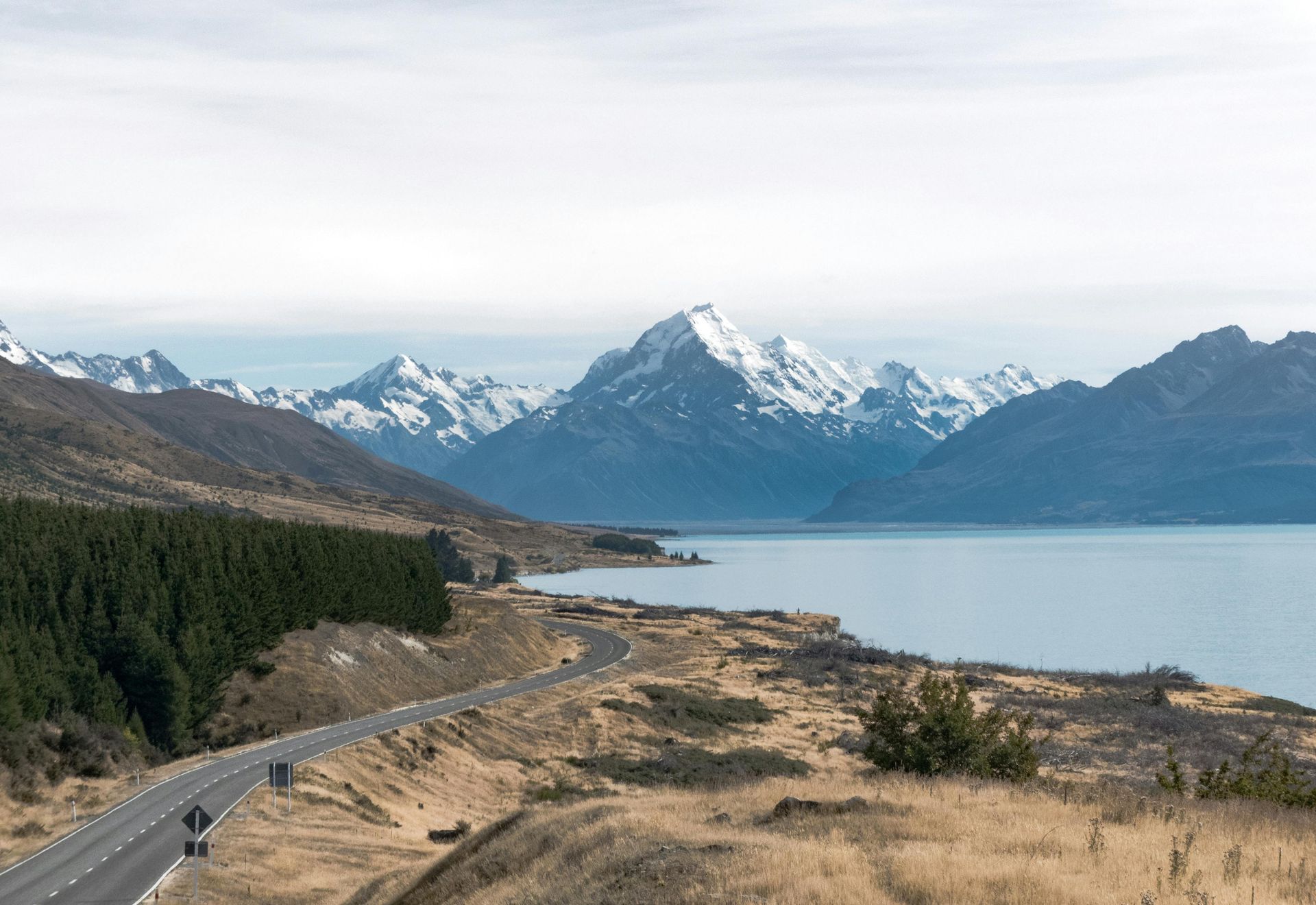 A car is driving down a road next to a lake with mountains in the background in New Zeland.