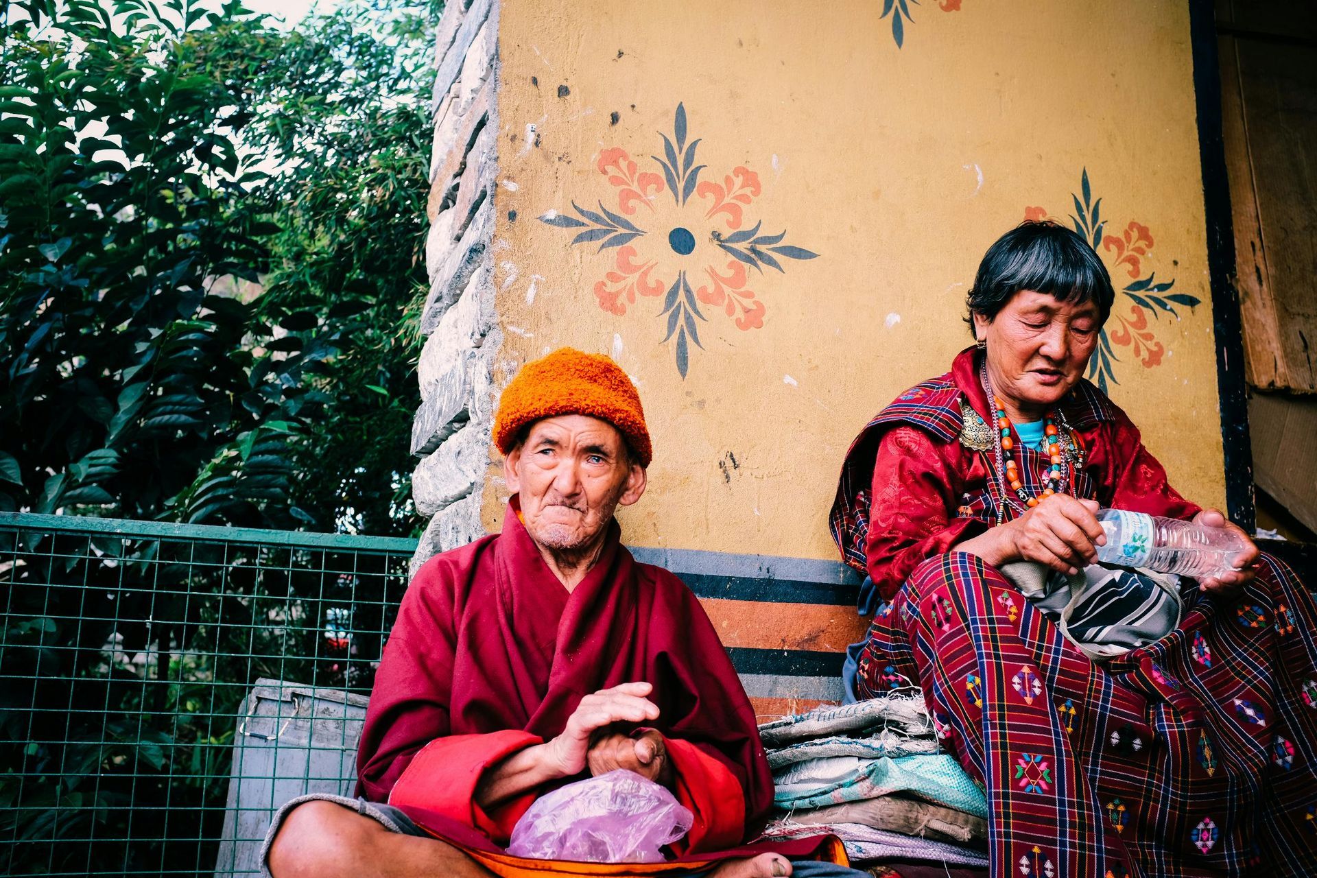 Two women are sitting on a bench in front of a yellow wall in Bhutan. 