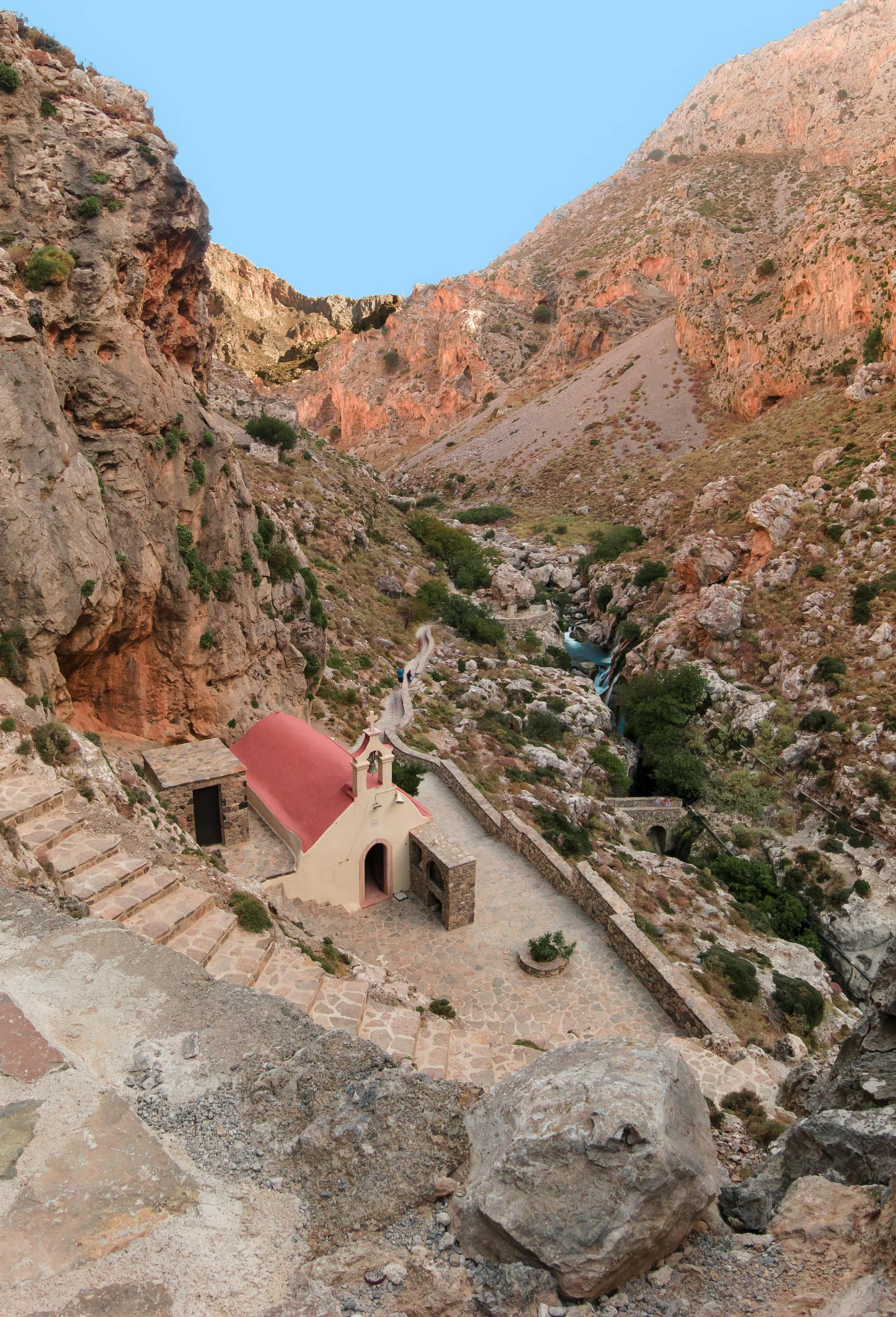 A small white building with a red roof is in the middle of a canyon in Crete, Greece.