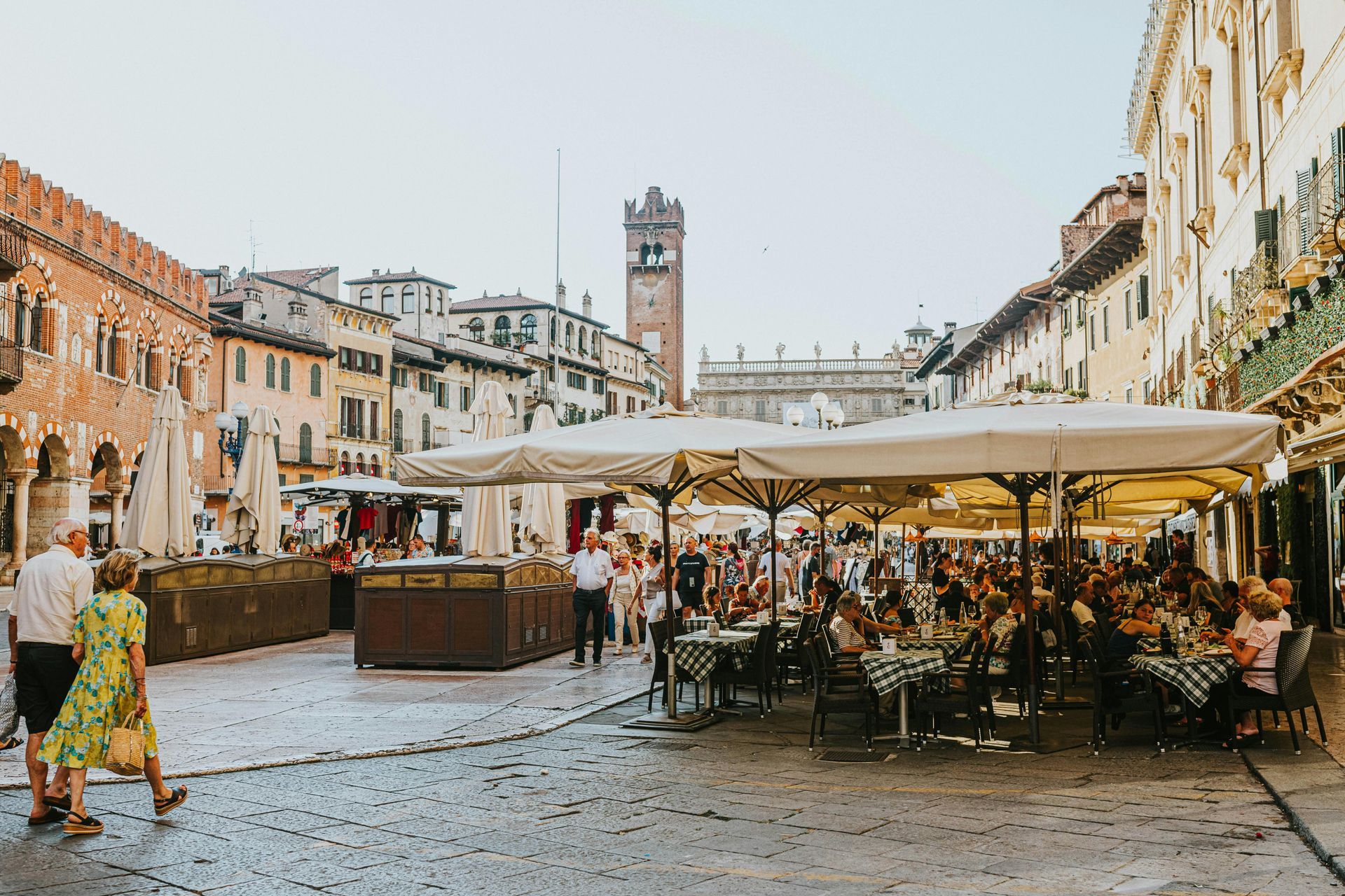 A group of people are sitting at tables under umbrellas in Verona, Italy's Square.