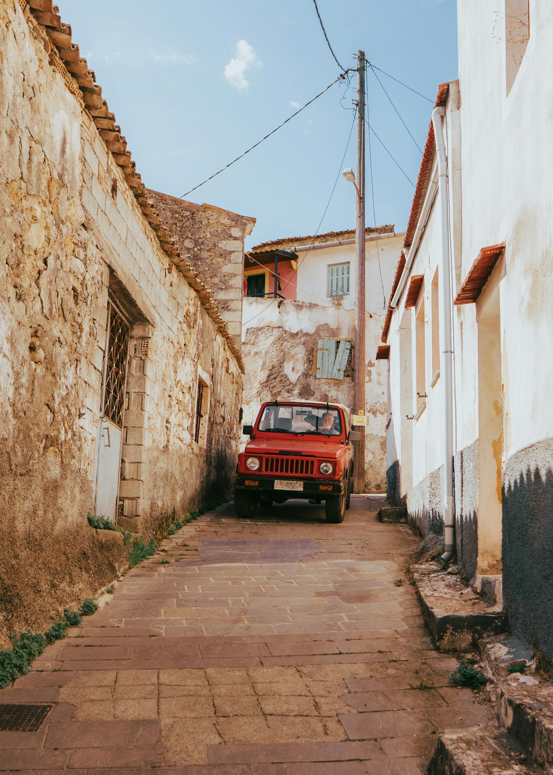 A red car is parked on the side of a narrow street in Corfu, Greece.