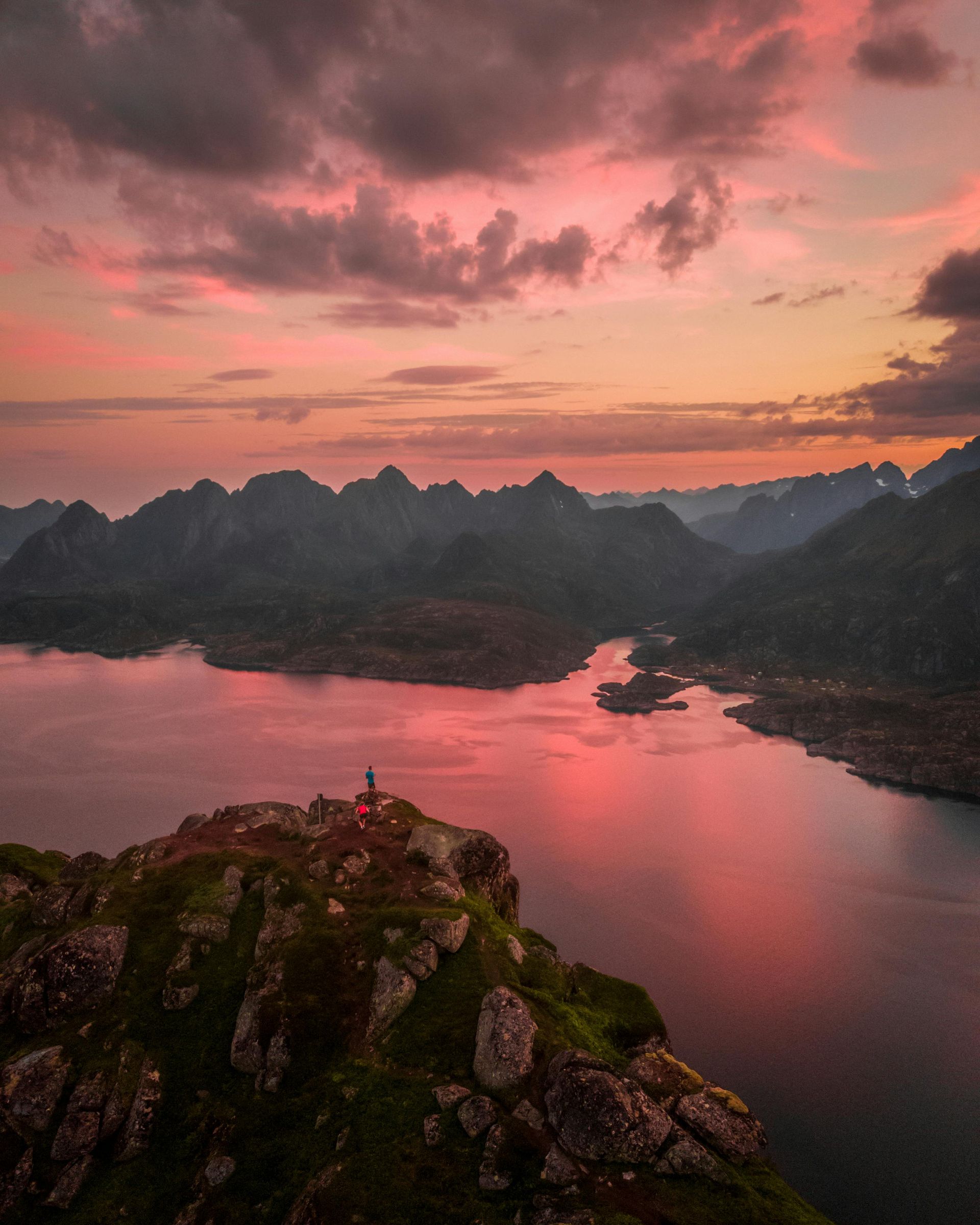 A person is standing on top of a mountain overlooking a lake at sunset on the Norwegian Coast.