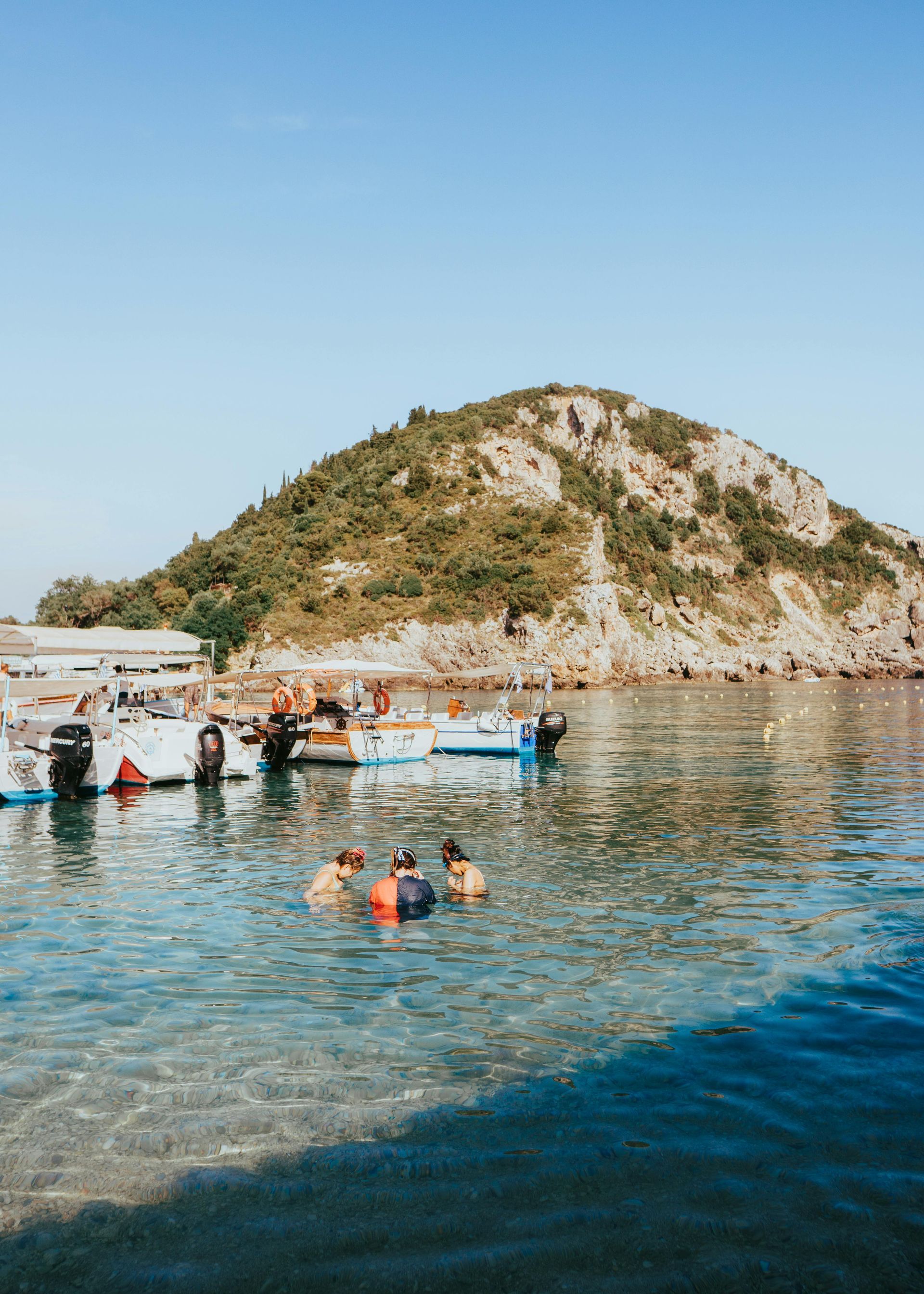 A group of people are swimming in the ocean with boats in the background in Corfu, Greece.