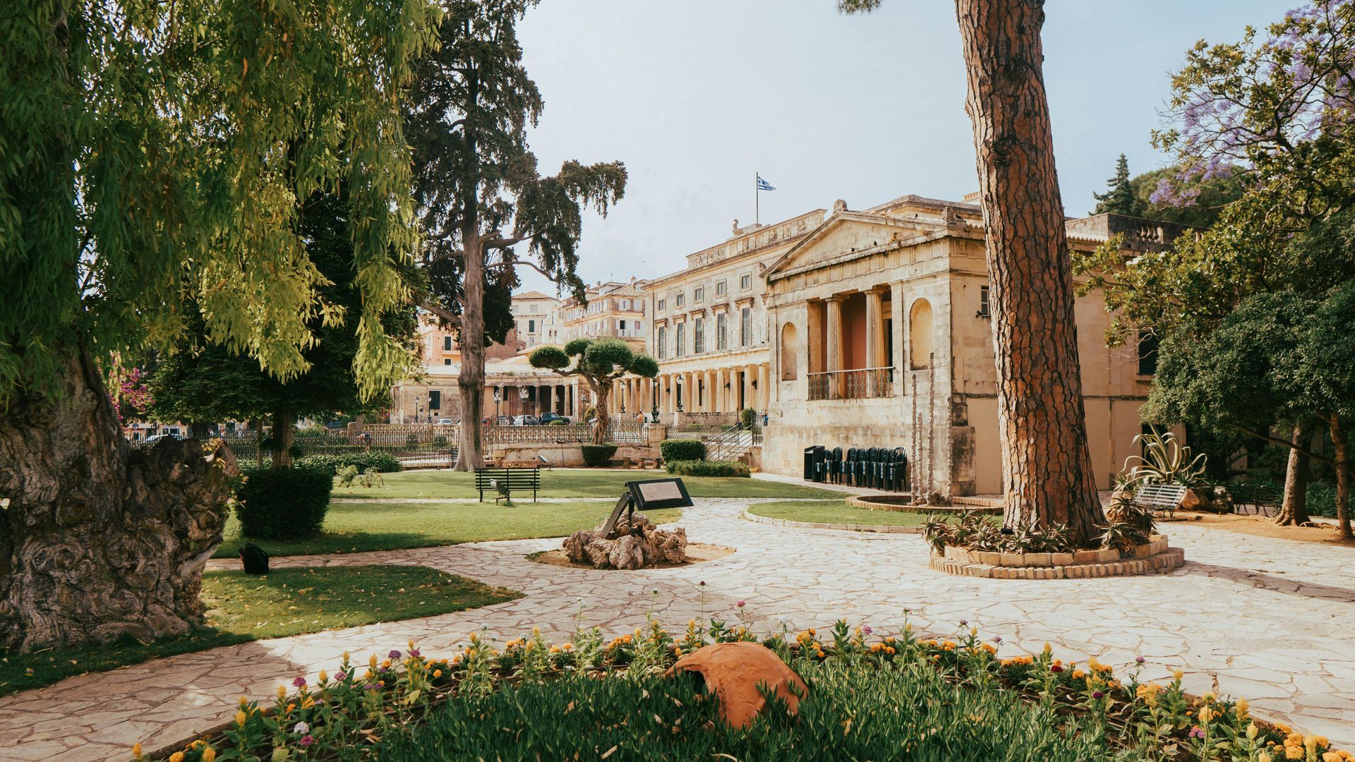 A large building is surrounded by trees and flowers in a park in Corfu, Greece.