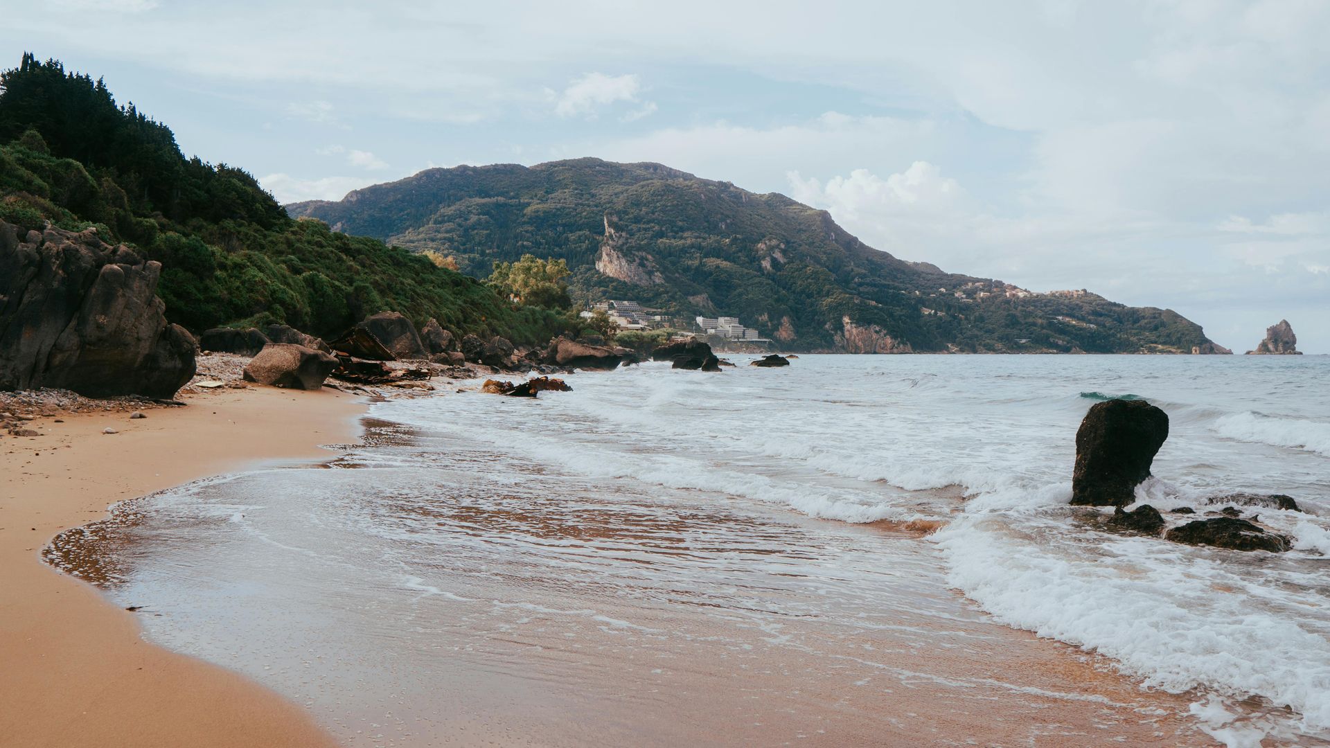 A beach with a mountain in the background and waves crashing on the sand in Corfu, Greece.