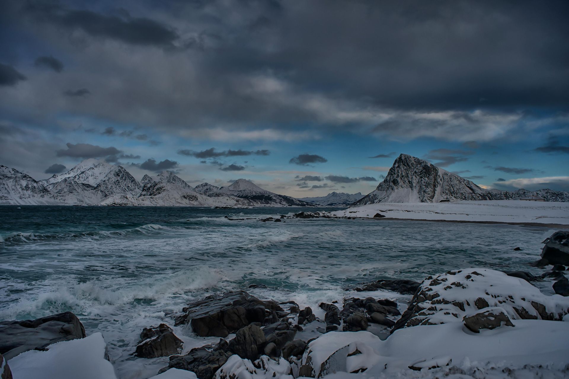 A snowy beach with mountains in the background and waves crashing on the rocks  on the Norwegian Coast.