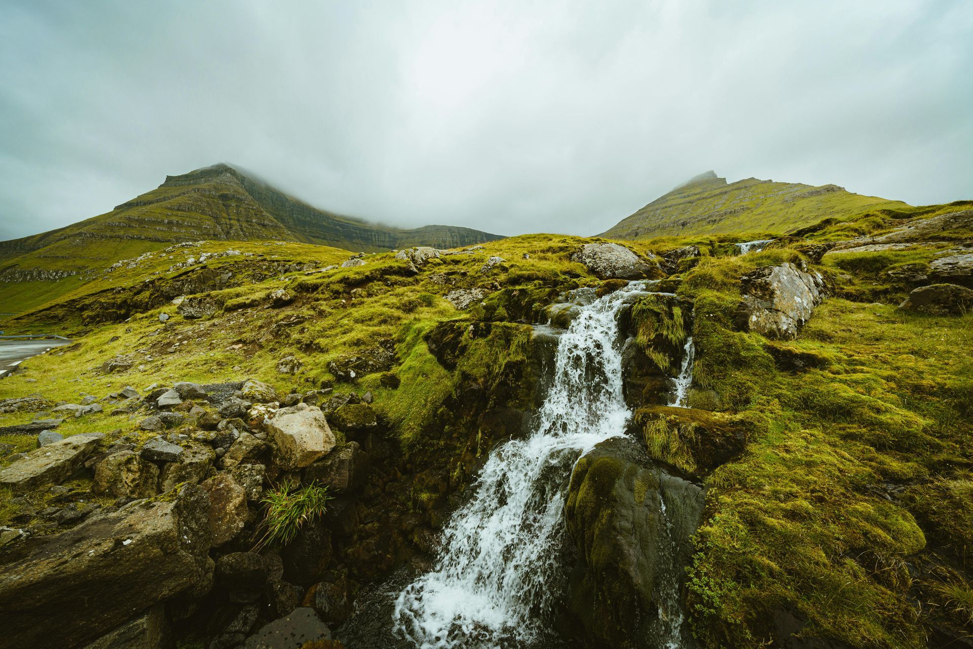 A small waterfall in the middle of a green field with mountains in the background on the Faroe Islands.