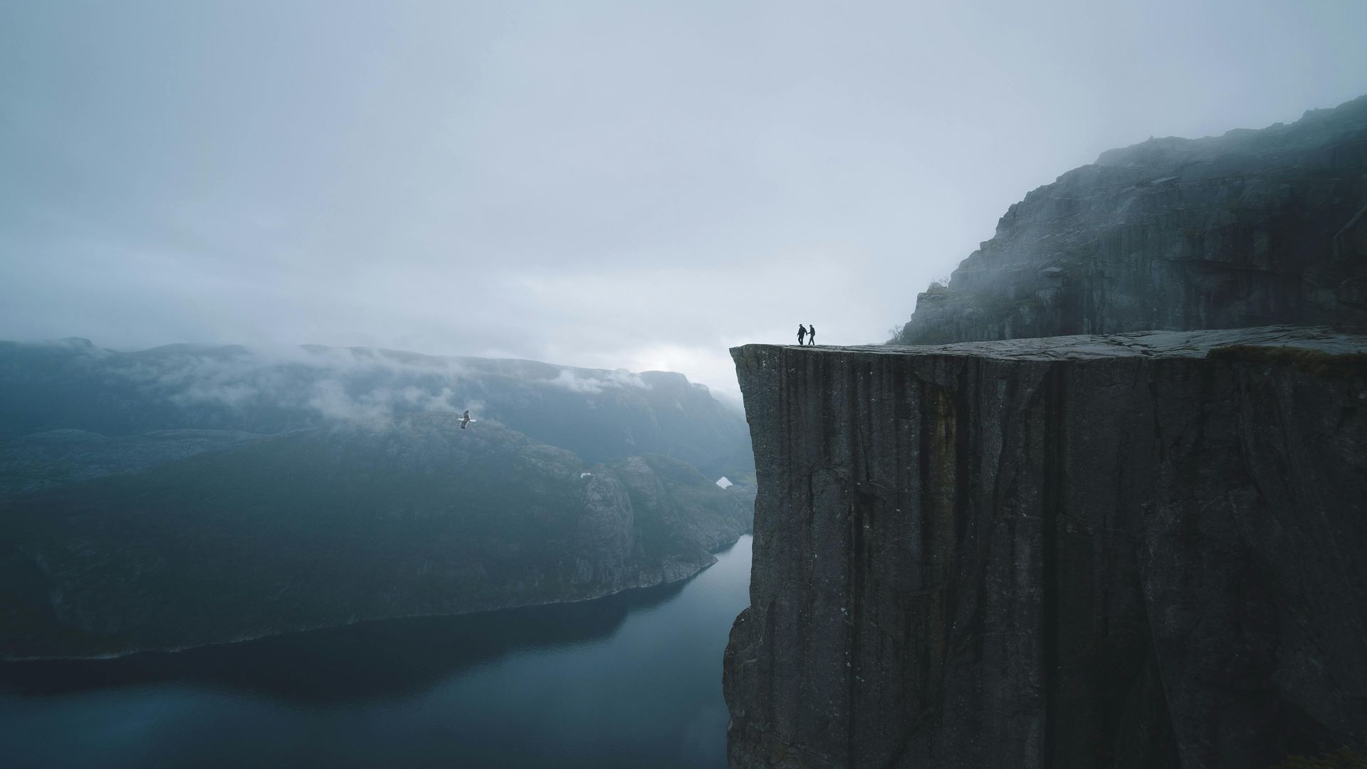 A couple is standing on the edge of a cliff overlooking a lake in Norway.