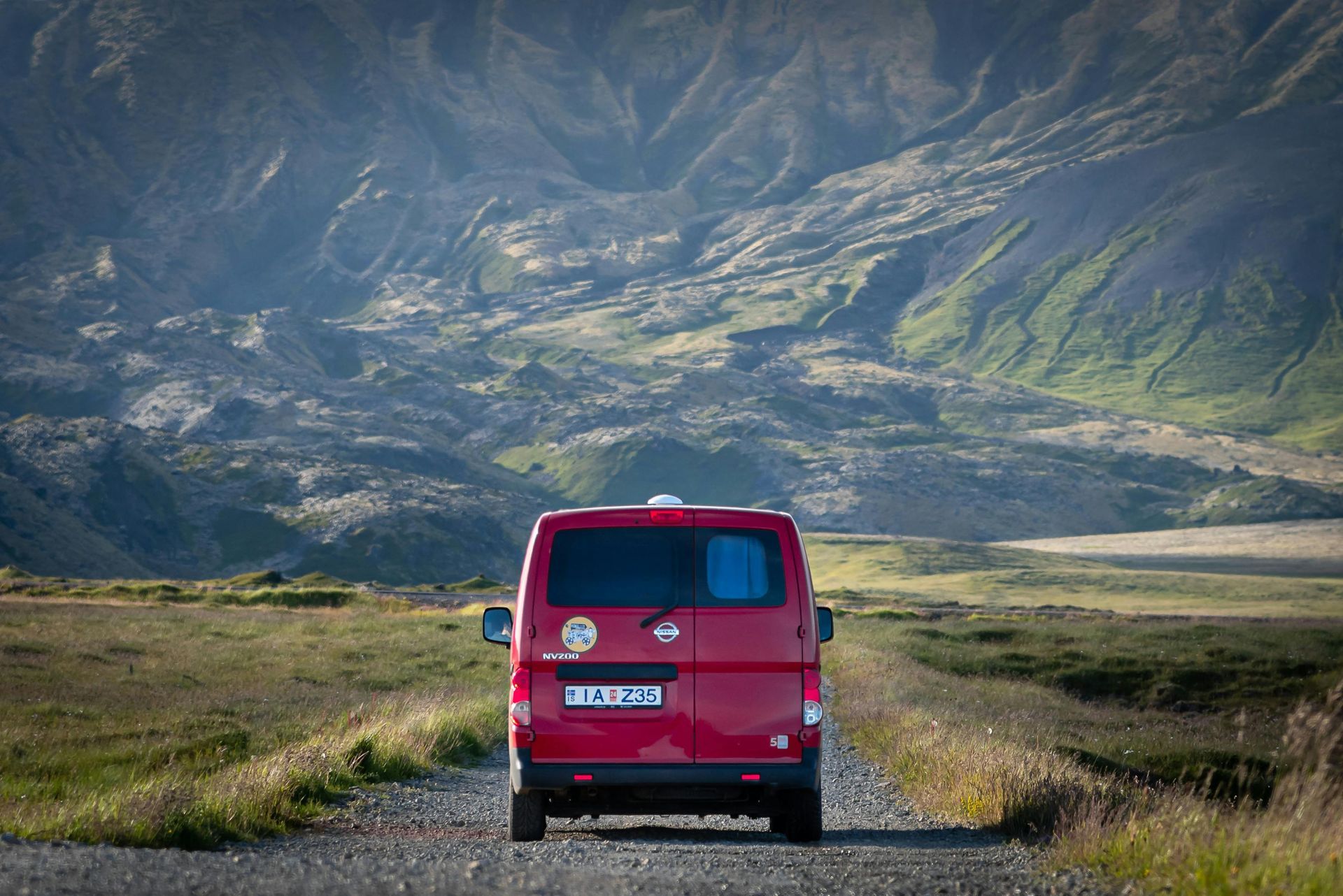 A red van is driving down a dirt road with mountains in the background in Iceland.