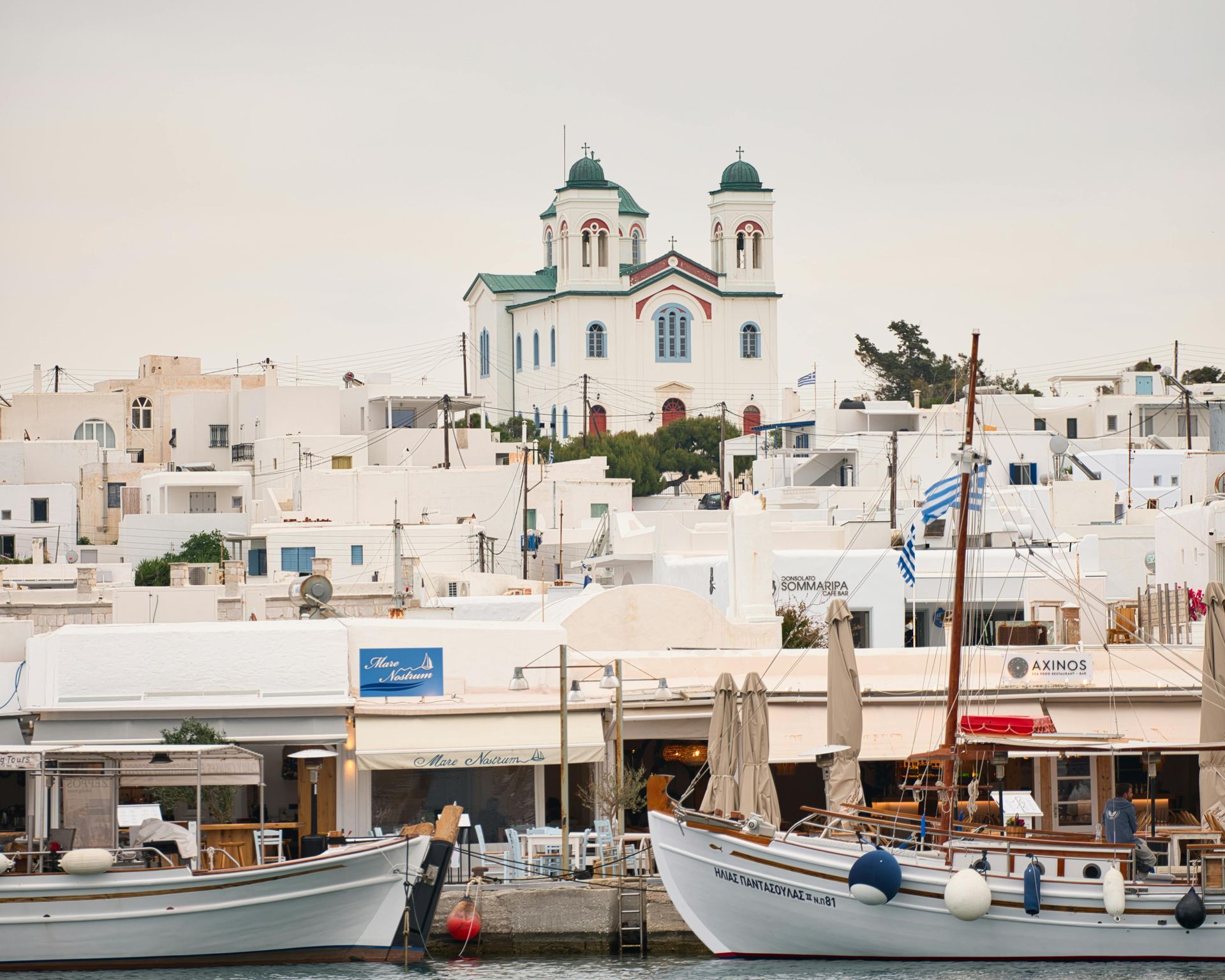 Boats are docked in a harbor with a church in the background in Paros, Greece.