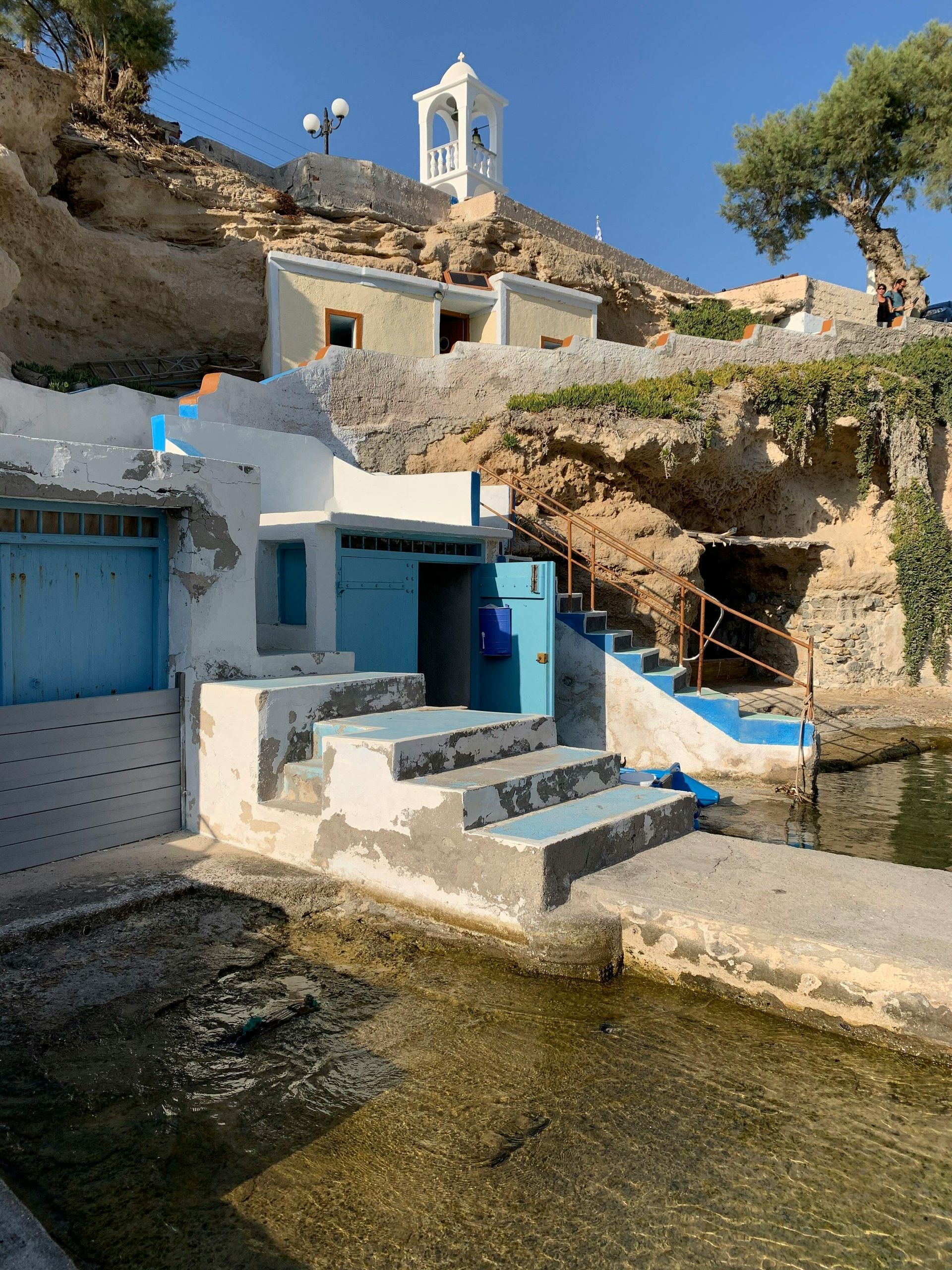 A blue and white building with stairs leading up to it next to a body of water in Milos, Greece.