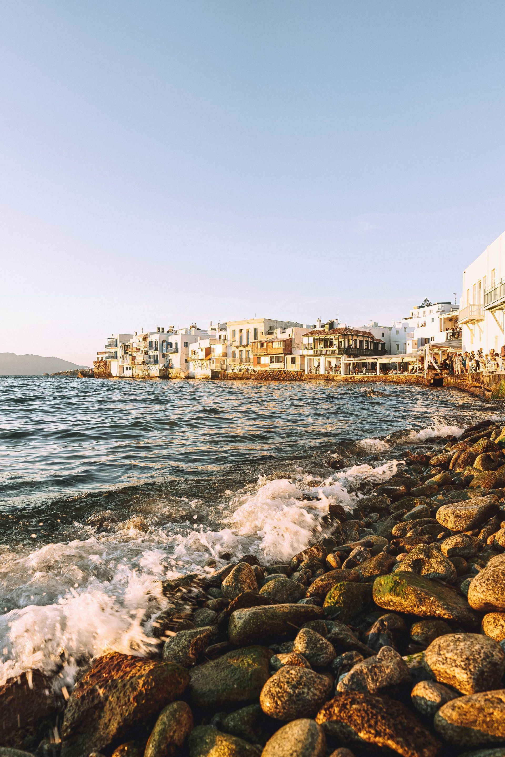 A rocky beach next to a body of water with buildings in the background in Mykonos, Greece.