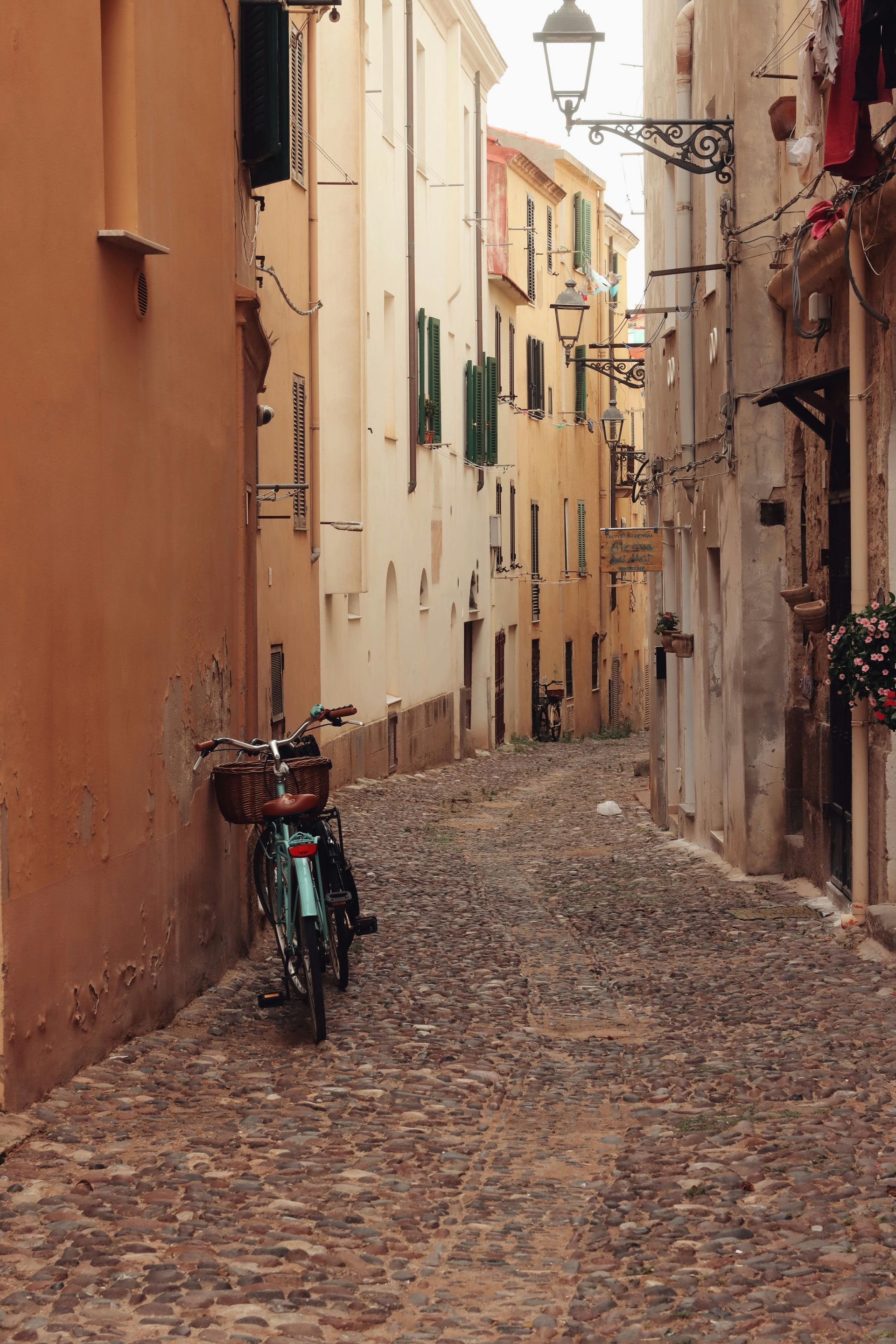 A motorcycle is parked on the side of a cobblestone street between two building in Sardinia, Italy.