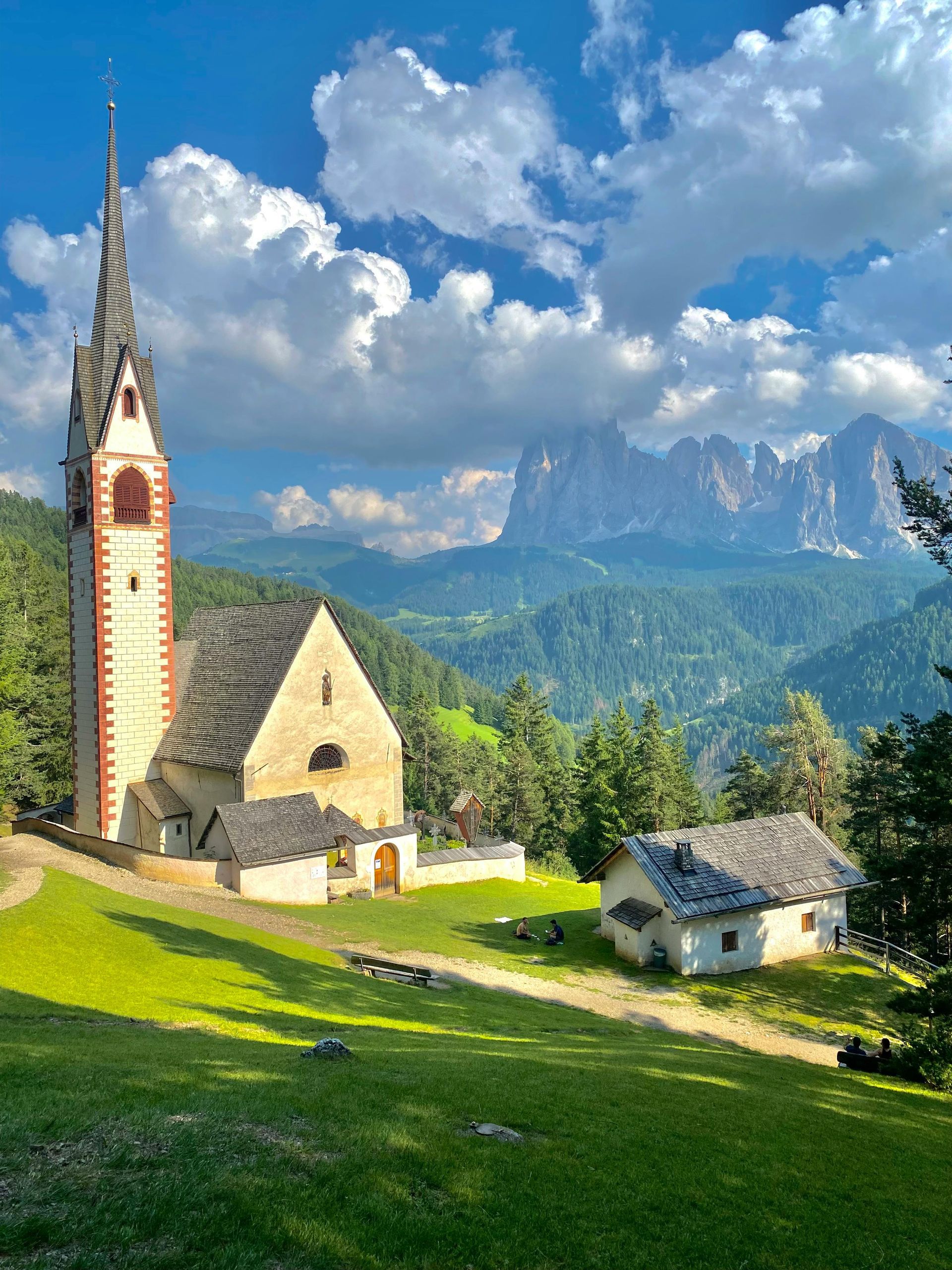 A church with a clock tower on top of a hill with The Dolomites in the background in Italy.