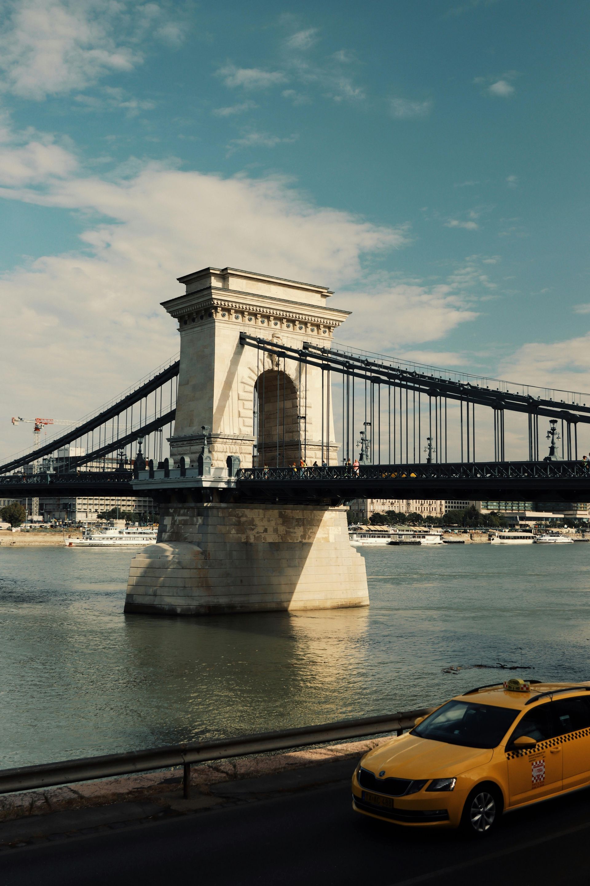 A yellow taxi is driving under a bridge over The Danube River.