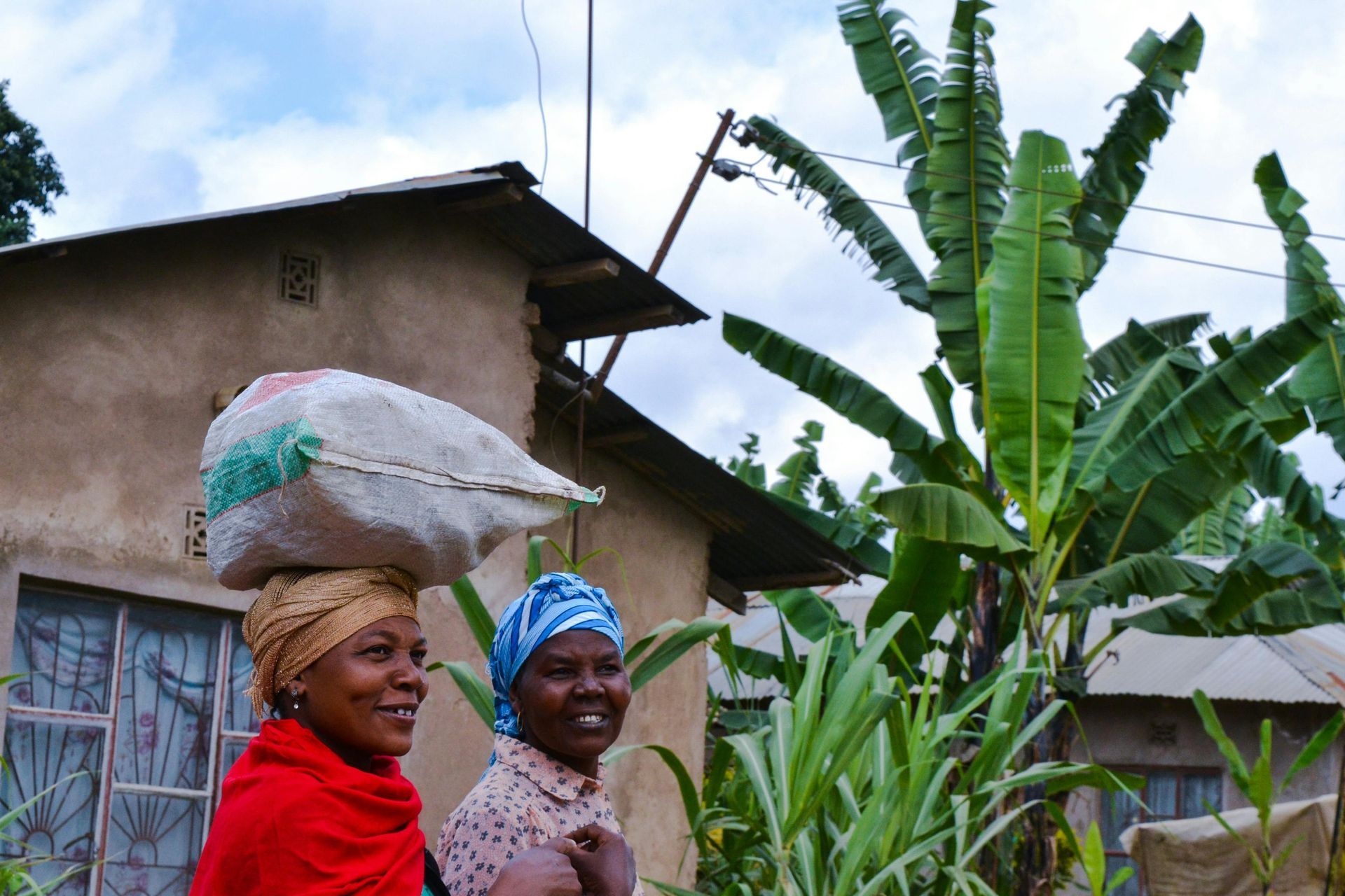 Two women are standing in front of a house with one carrying a large bag on her head in Tanzania, Africa.