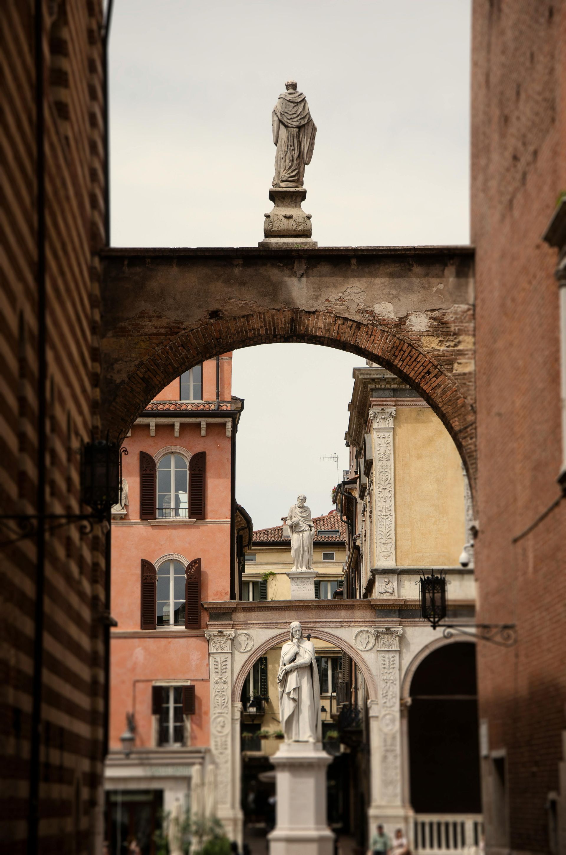 A statue of a man on top of an archway between two buildings in Verona, Italy.