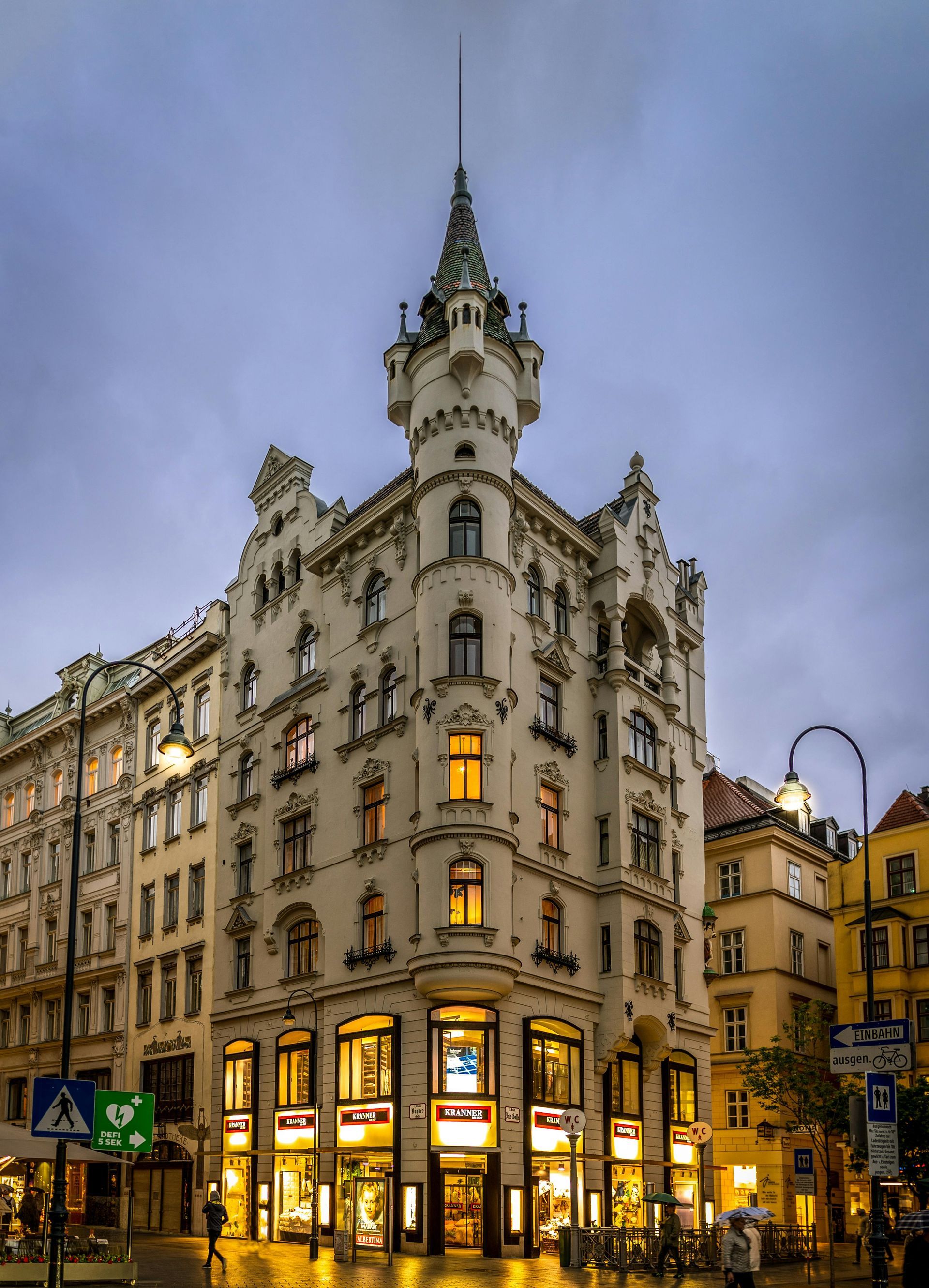 A large building with a tower on top of it is on the corner of a city street in Vienna.