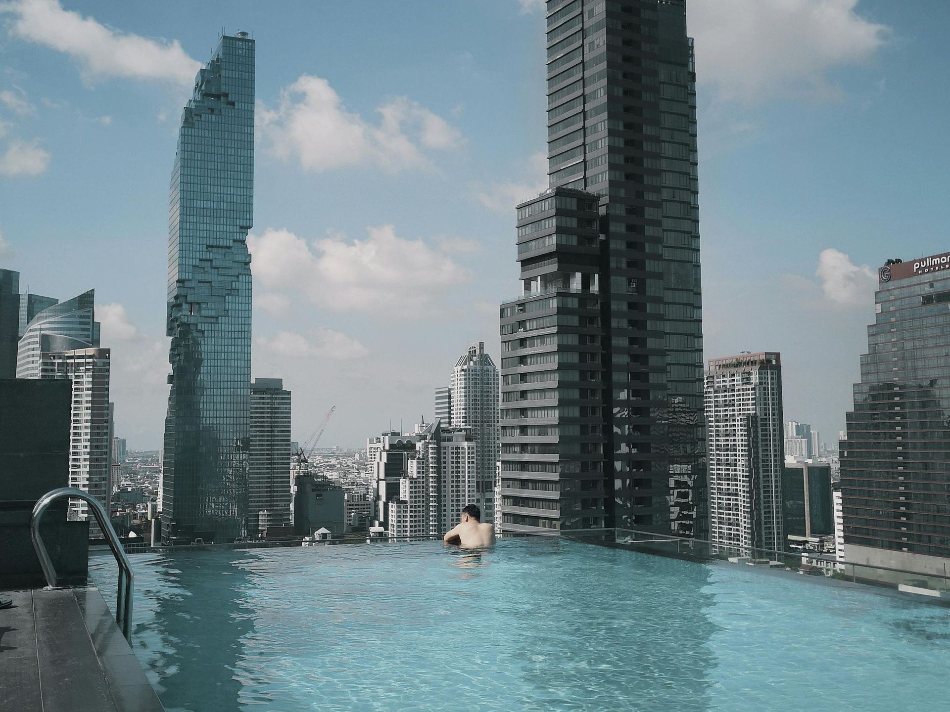 A man is swimming in an infinity pool with tall buildings in the background overlooking Bangkok, Thailand. 