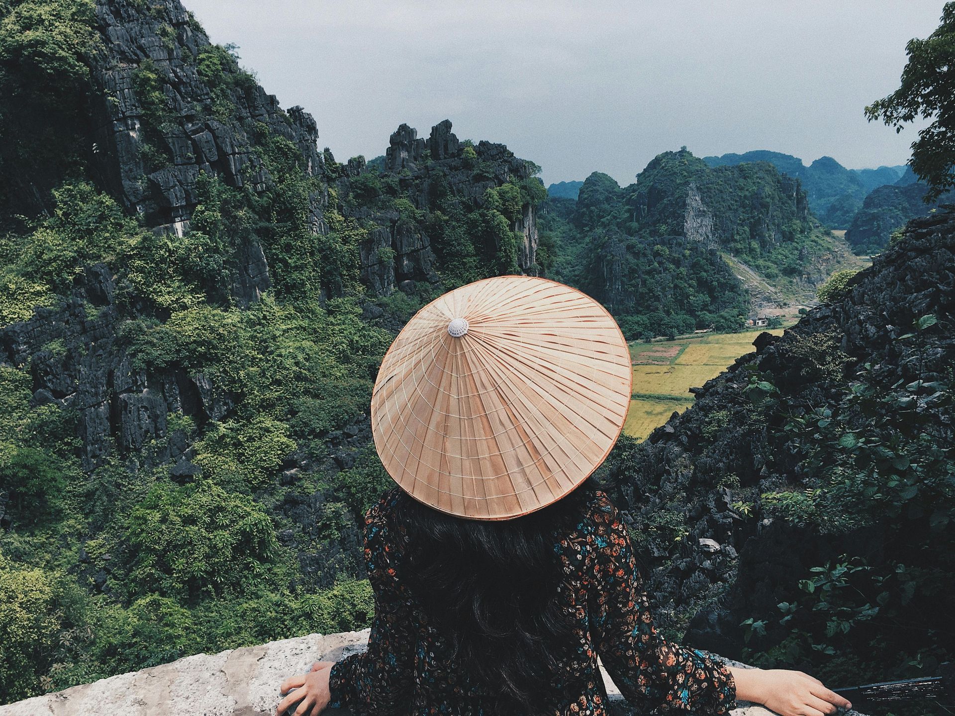 A woman wearing a conical hat is looking out over a mountain range in Vietnam.