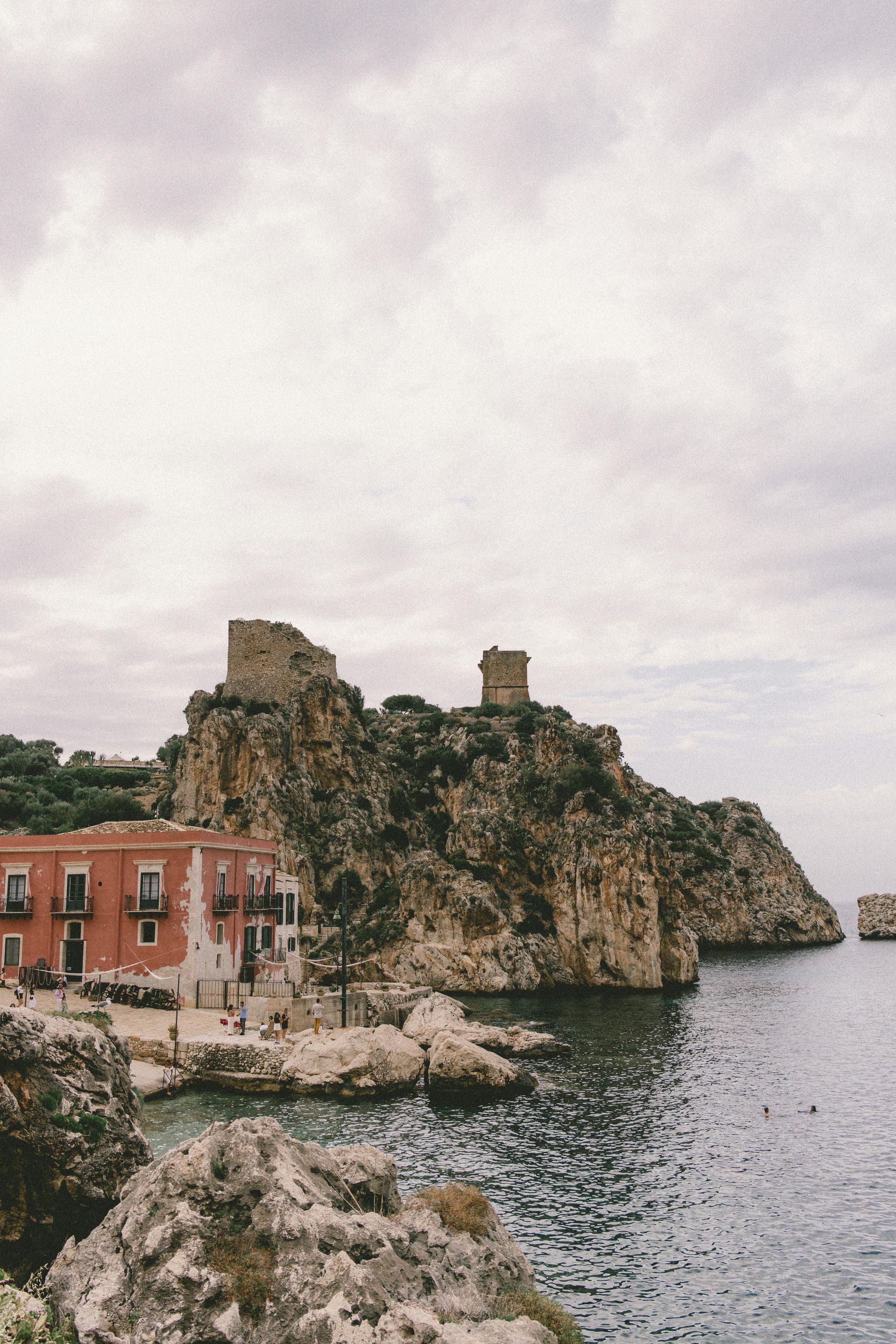 A castle sits on top of a rocky hill overlooking a body of water in Sicily, Italy.