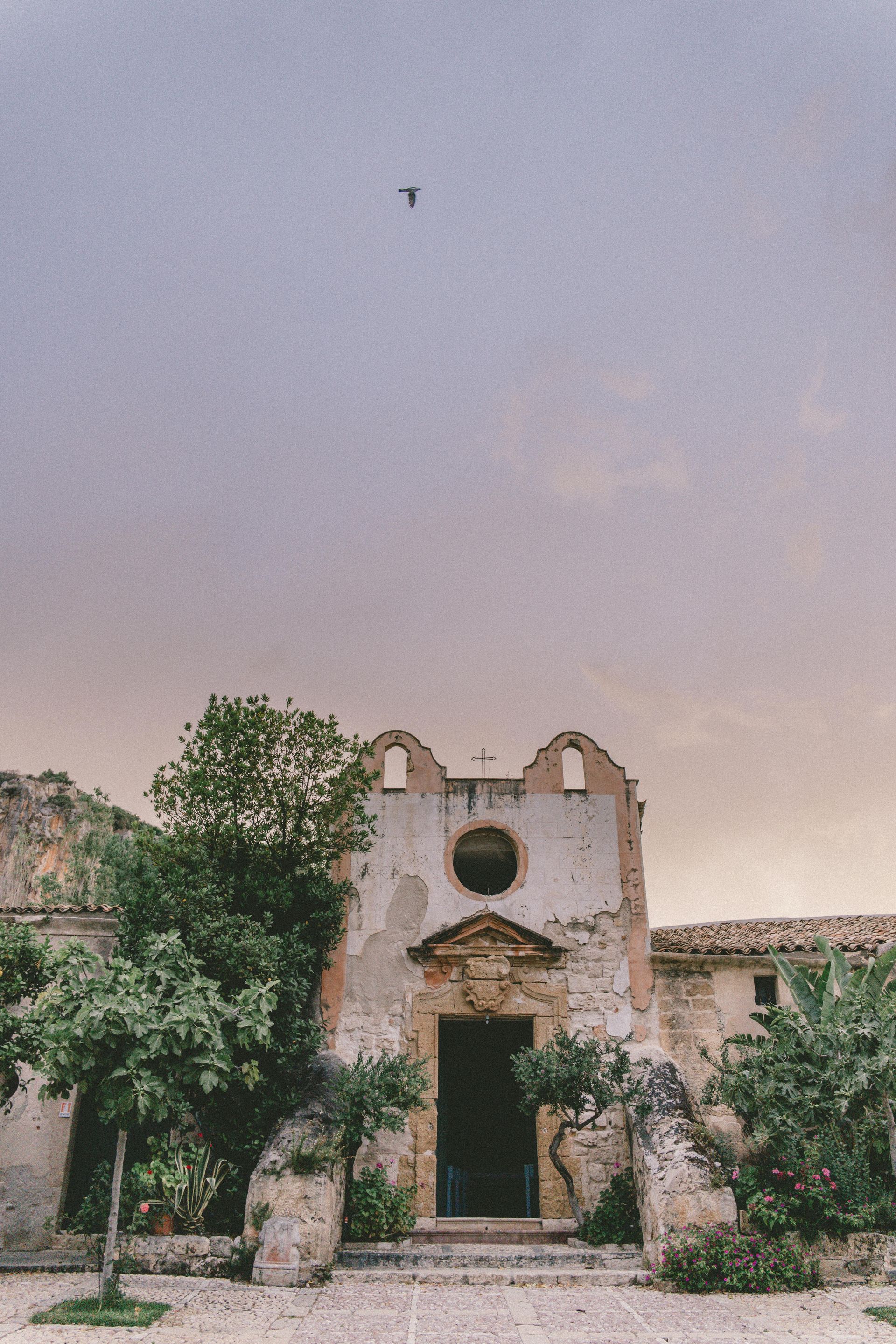 A bird is flying over an old building with trees in front of it in Sicily, Italy.