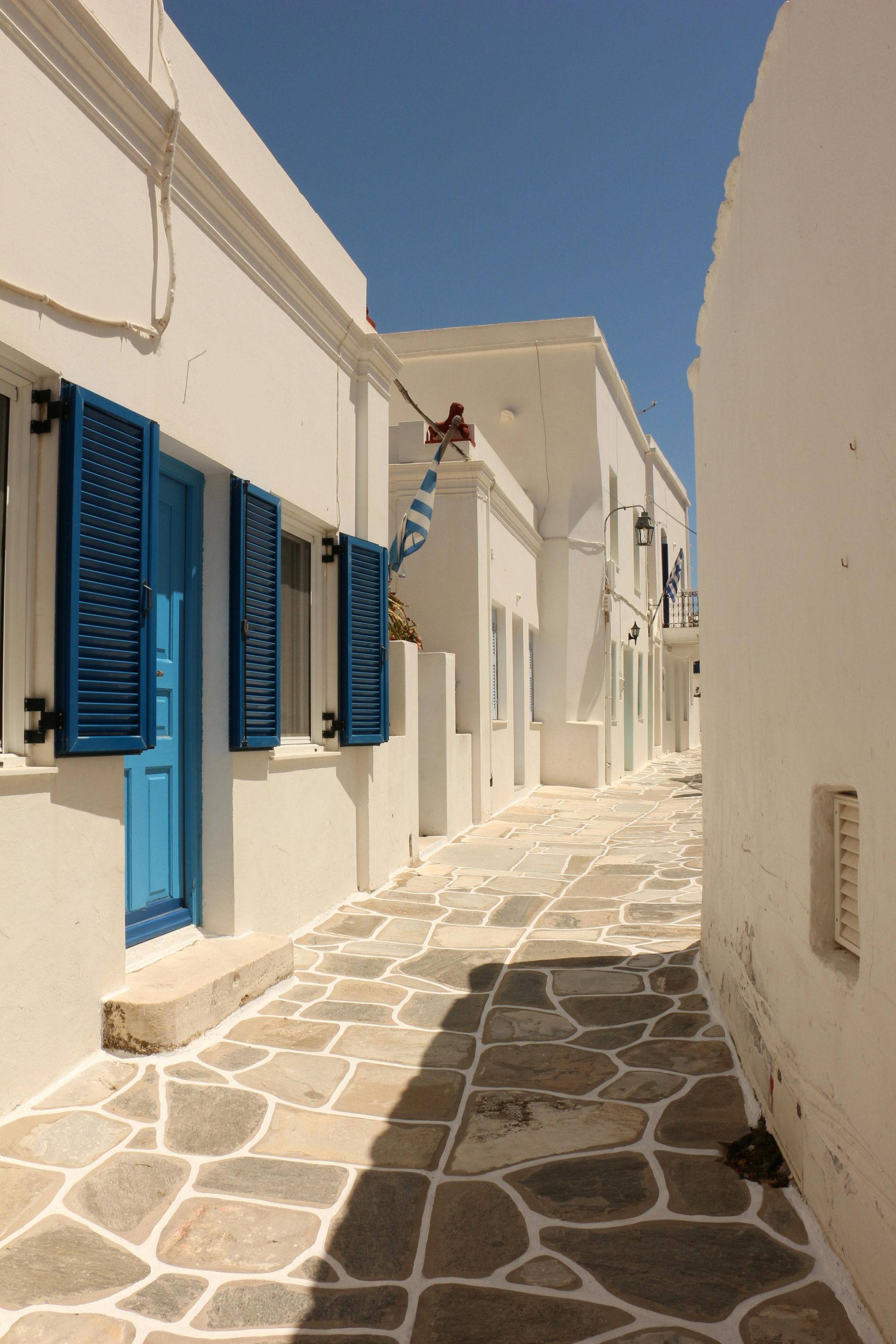 A narrow alleyway between two white buildings with blue shutters in Paros, Greece.