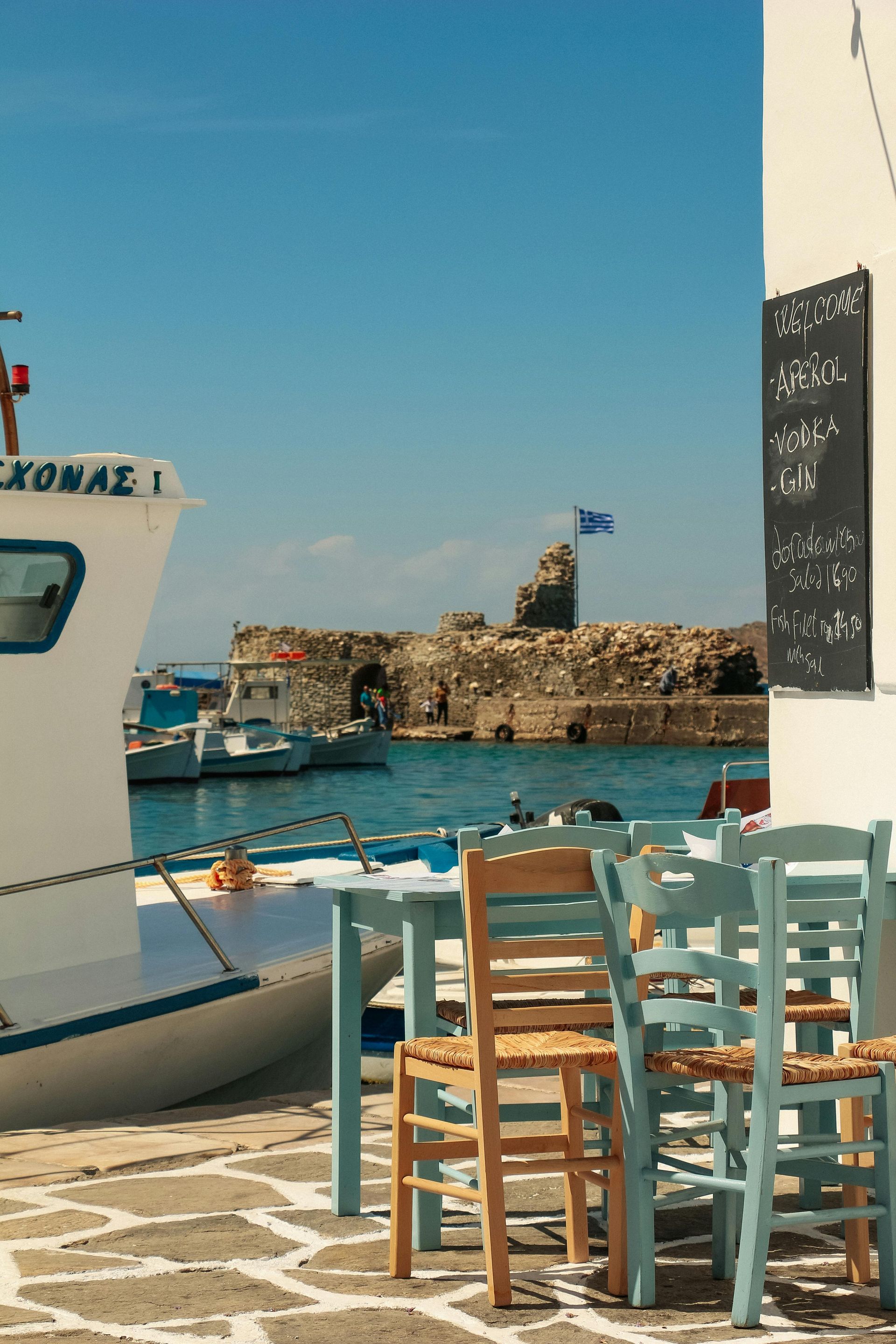 A boat is docked in a harbor with tables and chairs in front of it in Paros, Greece.